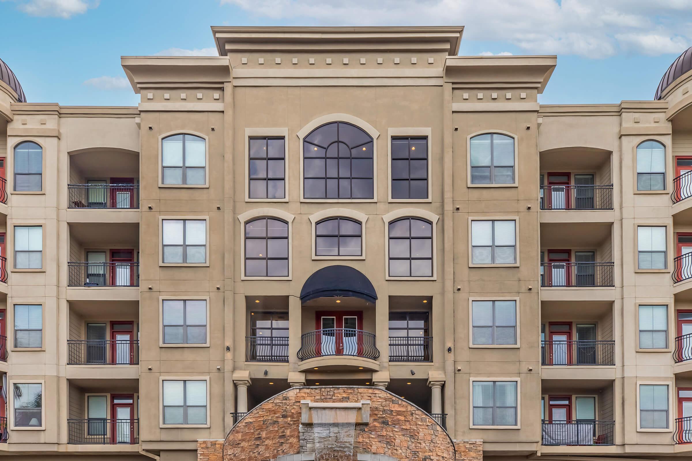 A modern multi-story apartment building featuring a symmetrical façade with large arched windows and balconies. The entrance is highlighted by an elaborate stone archway, and the structure is painted in neutral tones, contributing to a contemporary architectural style.
