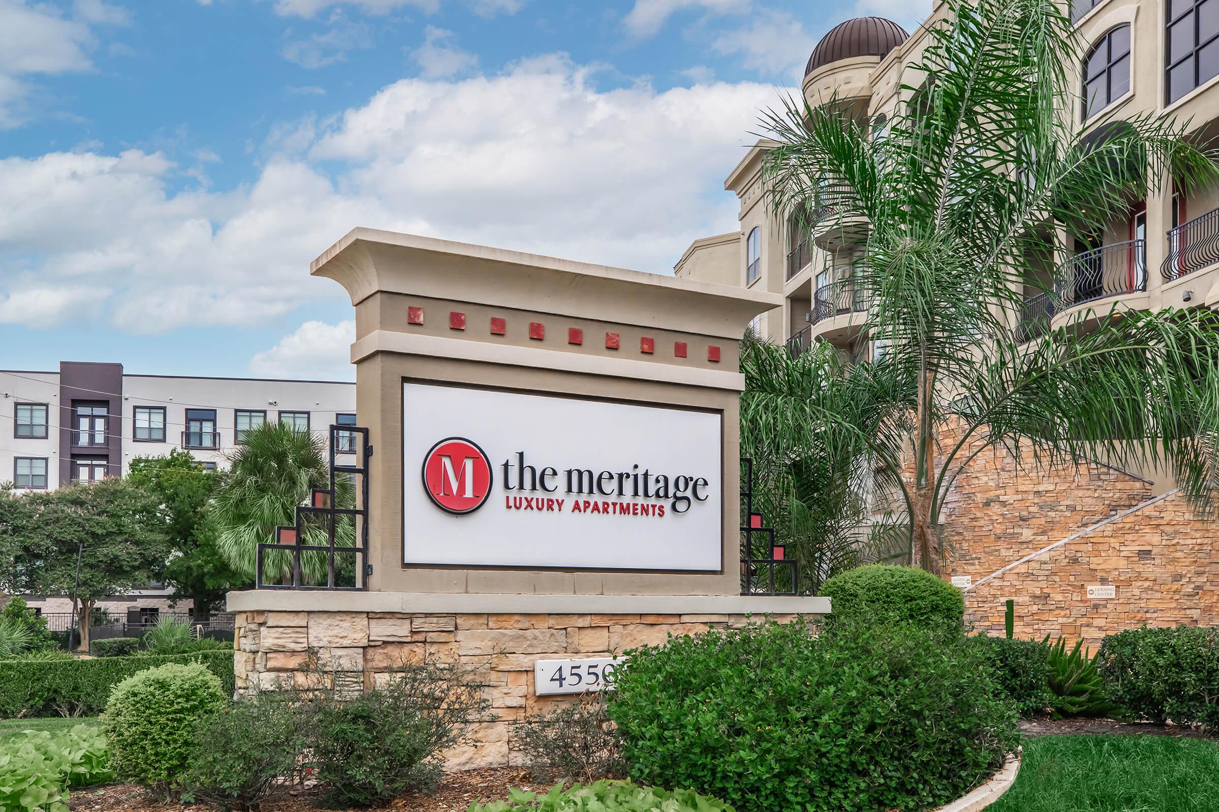Sign for The Meritage Luxury Apartments, featuring the name prominently with a red "M" logo. Surrounding greenery and a partially cloudy sky are visible, along with the address "4556" displayed on the sign. The setting appears to be a well-maintained apartment complex.