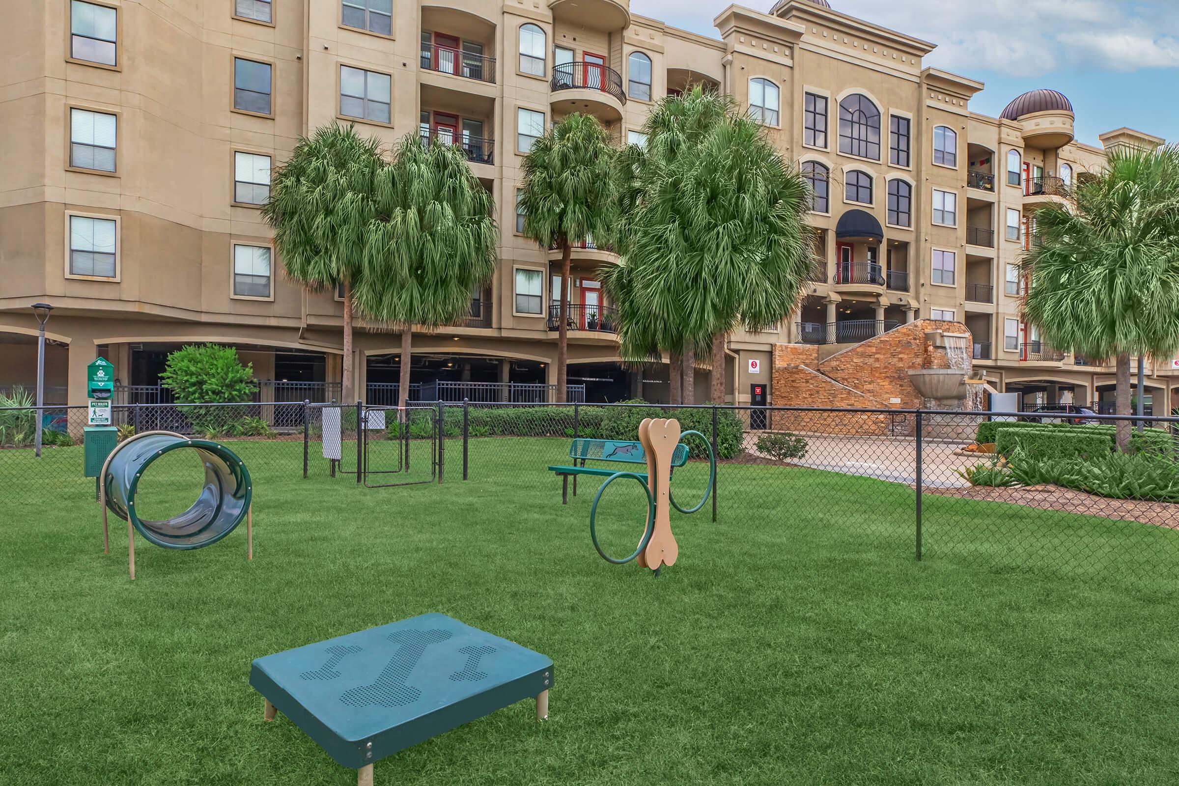 A well-maintained dog park with green grass, featuring various play structures like a tunnel, agility course, and benches. Surrounding the park are tall palm trees and a fenced area. In the background, a multi-story apartment building with balconies can be seen.