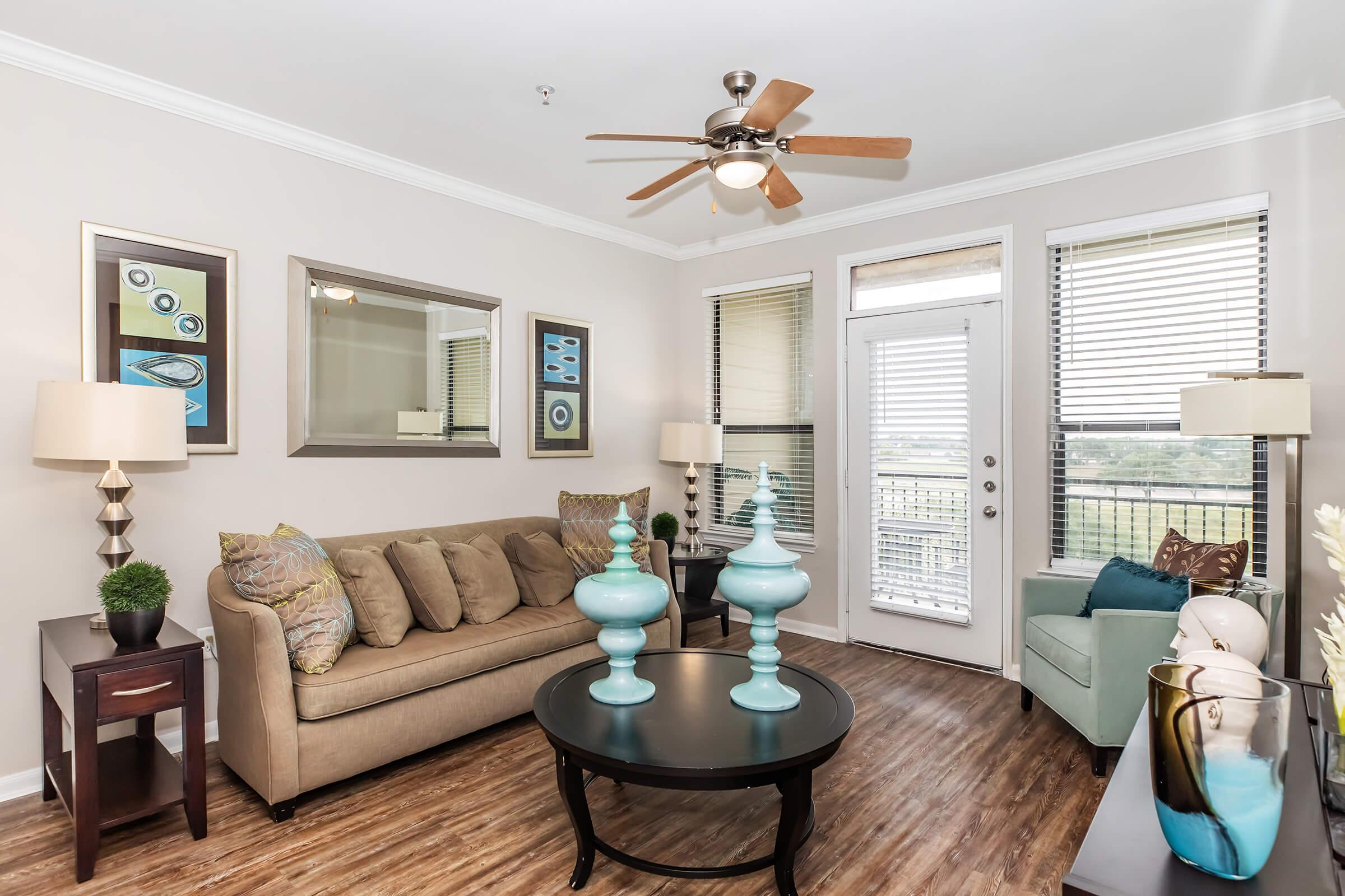 A cozy living room featuring a comfortable beige sofa, two decorative light blue vases on a round black coffee table, and an accent chair. There are tall lamps beside the sofa, framed artwork on the walls, and a large mirror reflecting natural light from windows with blinds. A door leads to an outdoor balcony area.