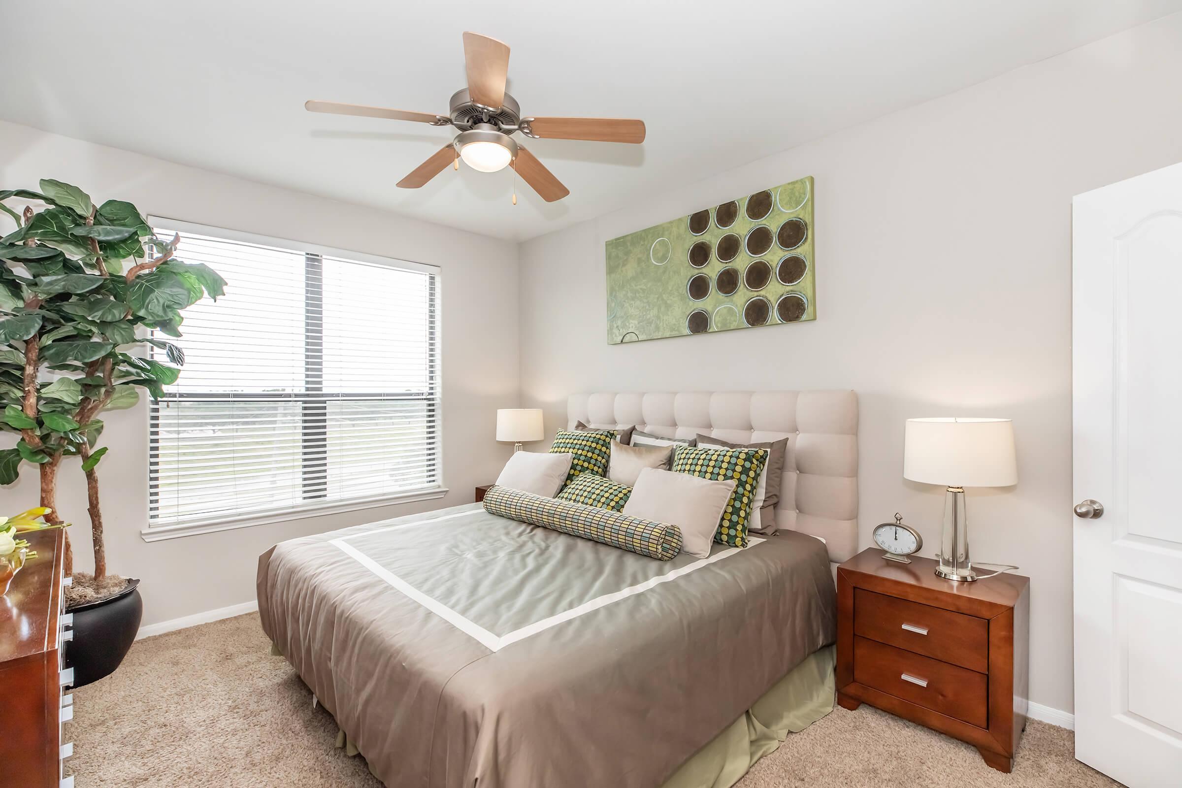 A cozy bedroom featuring a light-colored upholstered bed with decorative pillows, a wooden nightstand with a lamp, a potted plant in the corner, and a ceiling fan. Natural light filters through a window adorned with blinds, and abstract wall art complements the neutral tones of the decor.