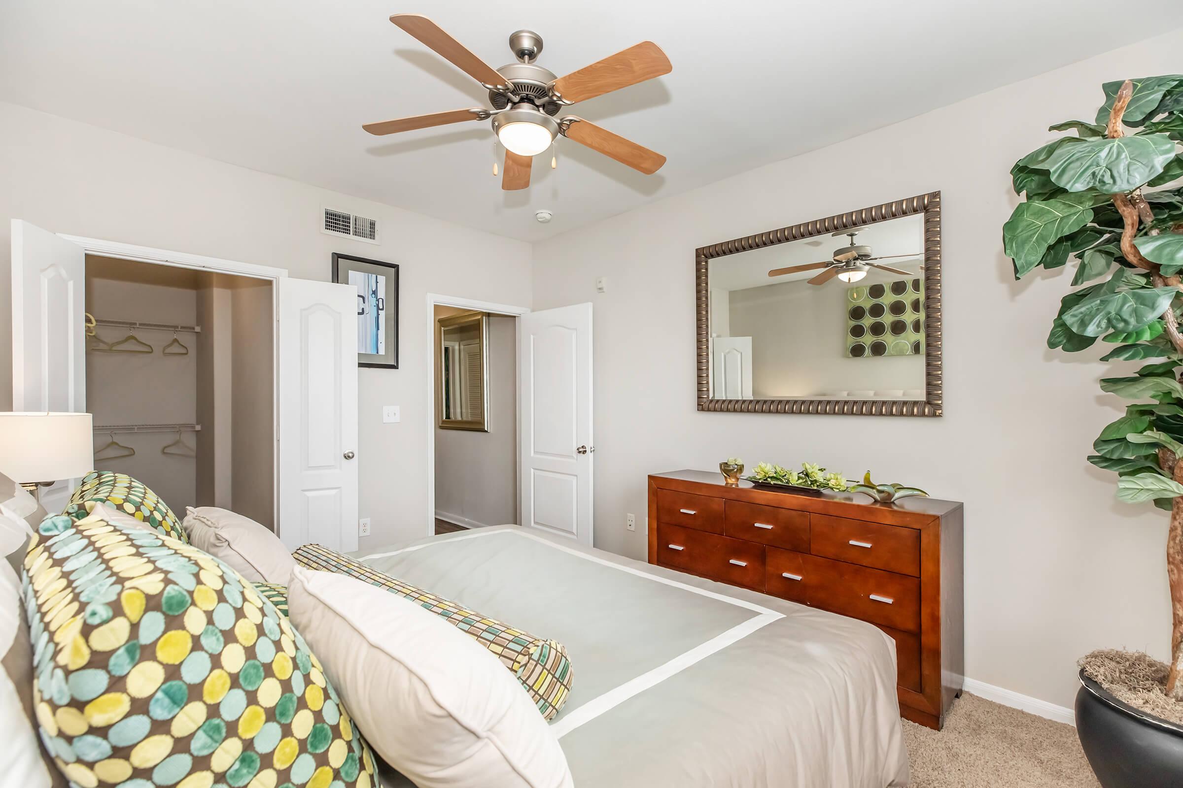 A cozy bedroom featuring a bed with decorative pillows, a wooden dresser, and a large mirror. A ceiling fan hangs above, and there are doors leading to a closet and another room. A potted plant adds a touch of greenery, enhancing the room's inviting atmosphere. Walls are painted in neutral tones.
