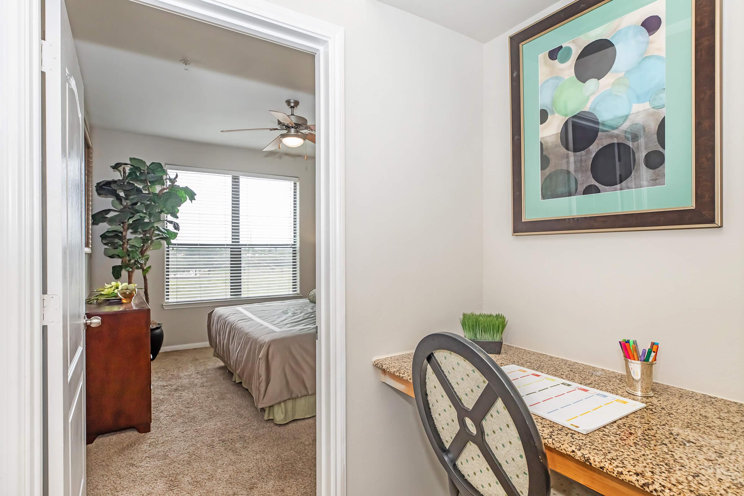 A view from a small workspace with a desk, colorful pens, and a plant, leading into a cozy bedroom. Natural light filters through a window with blinds, illuminating the neutral-toned decor. A framed abstract artwork hangs on the wall, adding a pop of color to the indoor space.