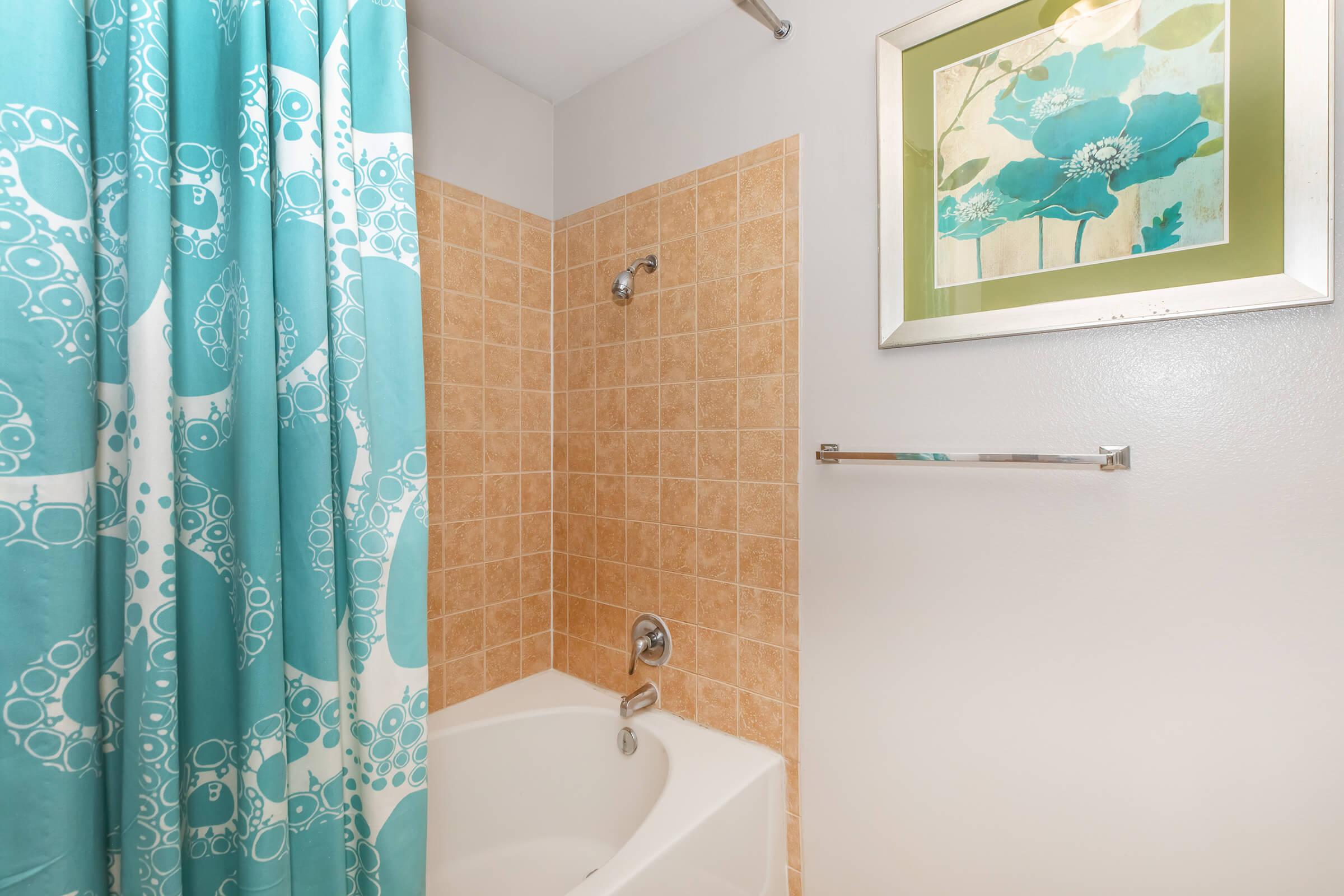 A bathroom featuring a bathtub with a shower, adorned with a turquoise and white patterned shower curtain. The tiled wall has a warm, beige color, and a framed green and blue floral artwork is mounted above the bathtub. A chrome towel bar is visible on the right side.