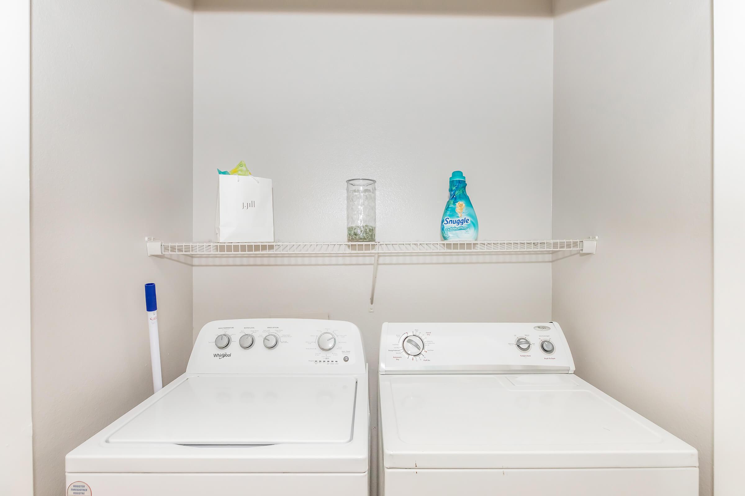 A laundry room featuring a white washing machine and dryer side by side. Above them is a shelf with a glass container and a bottle of laundry detergent. A blue cleaning tool is positioned on the left side. The walls are light-colored, providing a clean and organized appearance.