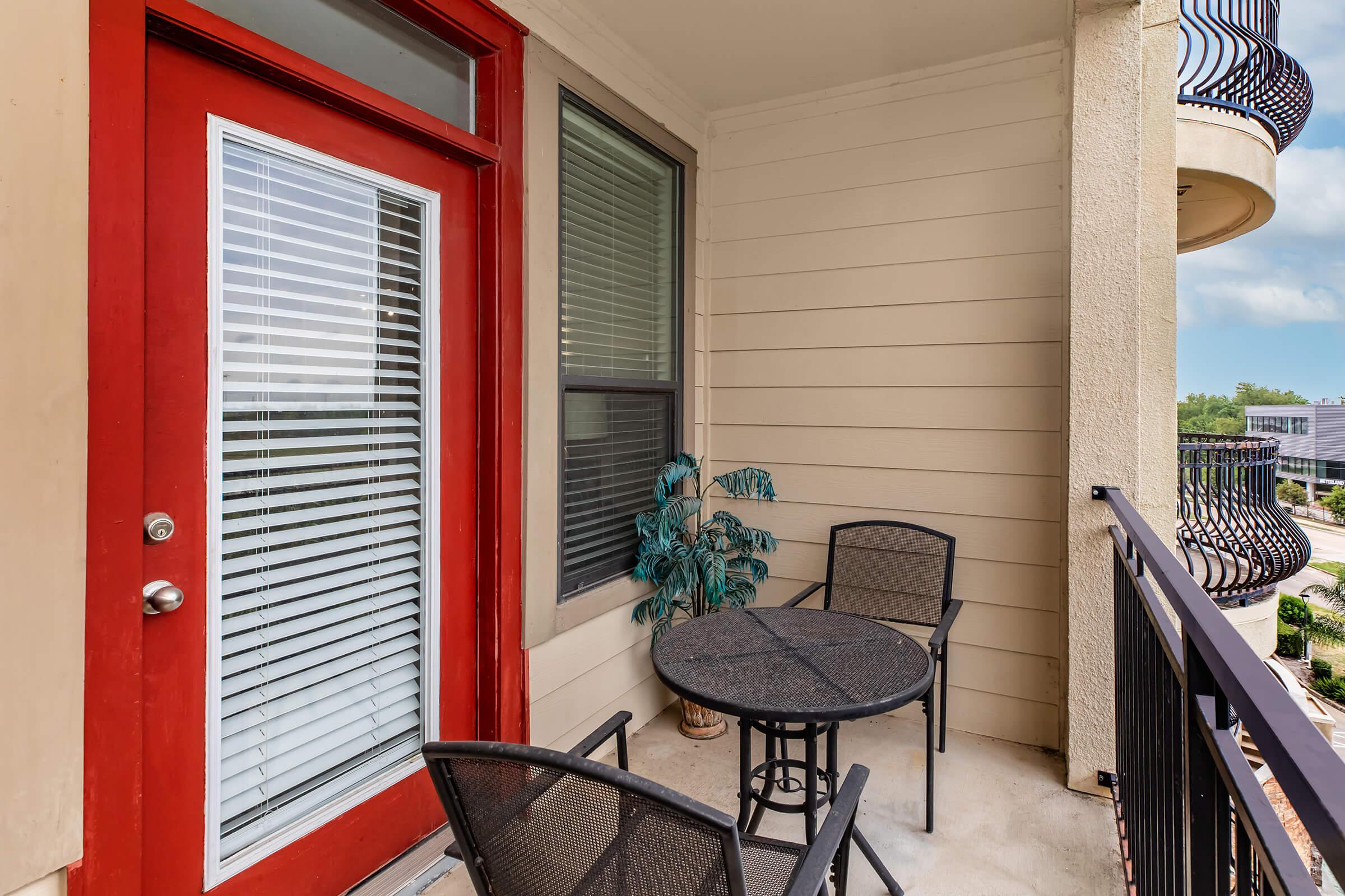A small balcony featuring a round black table and two chairs, with a green plant in a pot. The balcony is enclosed by a railing and has a door with vertical blinds leading into a room. The walls are light-colored, creating a cozy outdoor space.