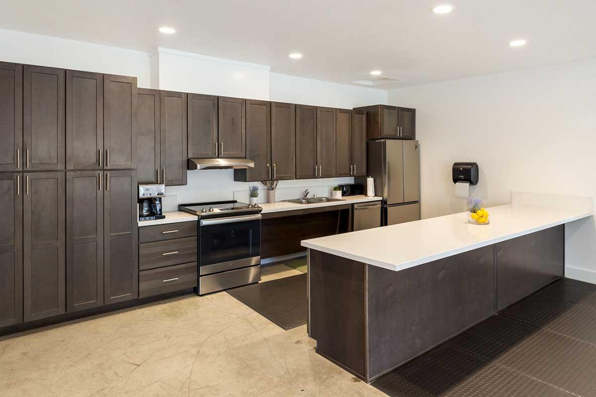 Modern kitchen featuring dark wood cabinets, a stainless steel refrigerator, and an oven. There is a white countertop island with a vase of lemons, a coffee maker, and various small appliances. Bright overhead lighting illuminates the space, which has a clean and contemporary design.
