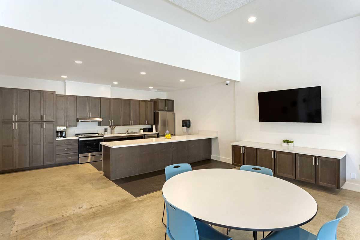 Modern kitchen and dining area featuring dark wood cabinetry, stainless steel appliances, a large central island, and a round table with blue chairs. Bright lighting and a large wall-mounted TV are also visible, creating a functional and inviting space for gatherings or communal cooking.