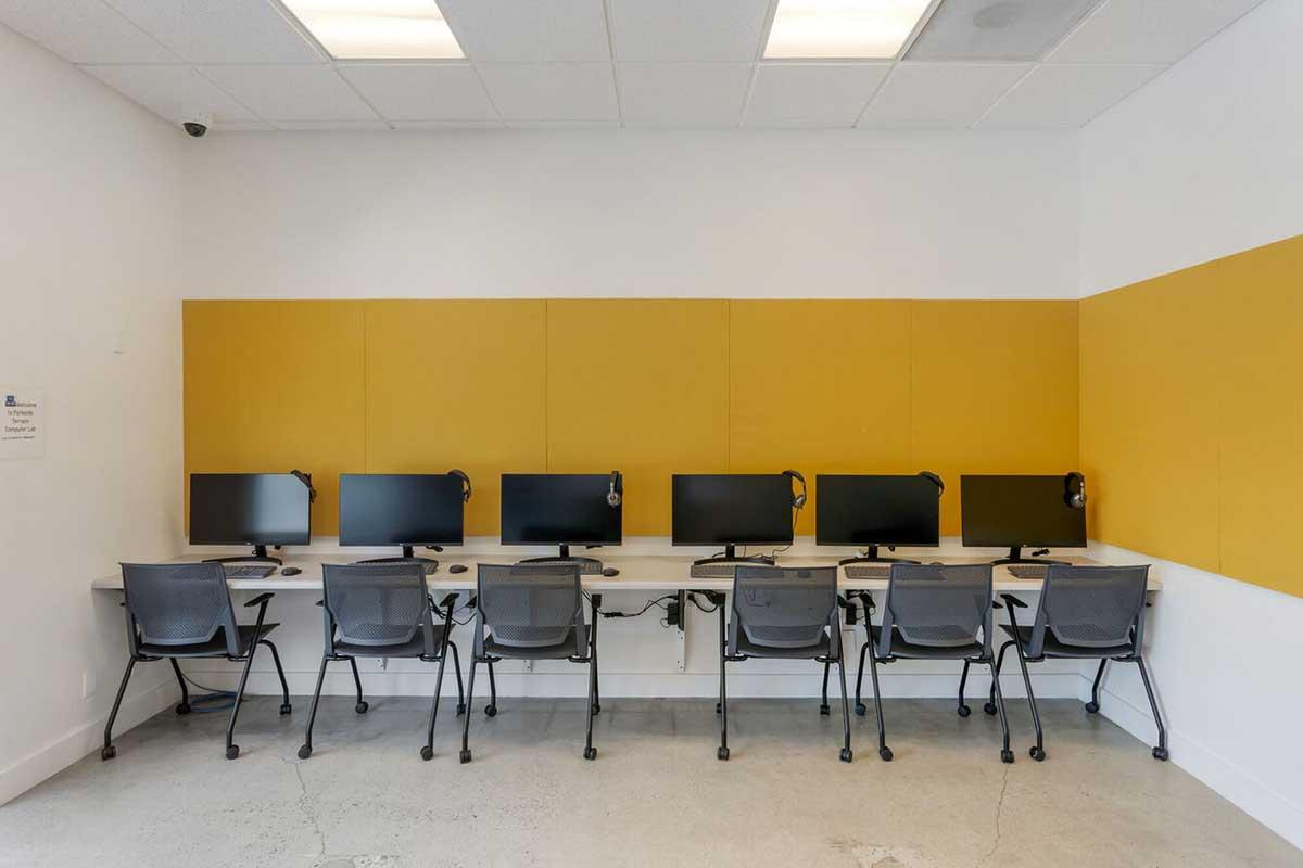 A row of six desktop computers with monitors and headsets, arranged on a long desk against a bright yellow wall. The room has a modern design, featuring neutral-colored flooring and overhead lighting, creating a functional workspace.