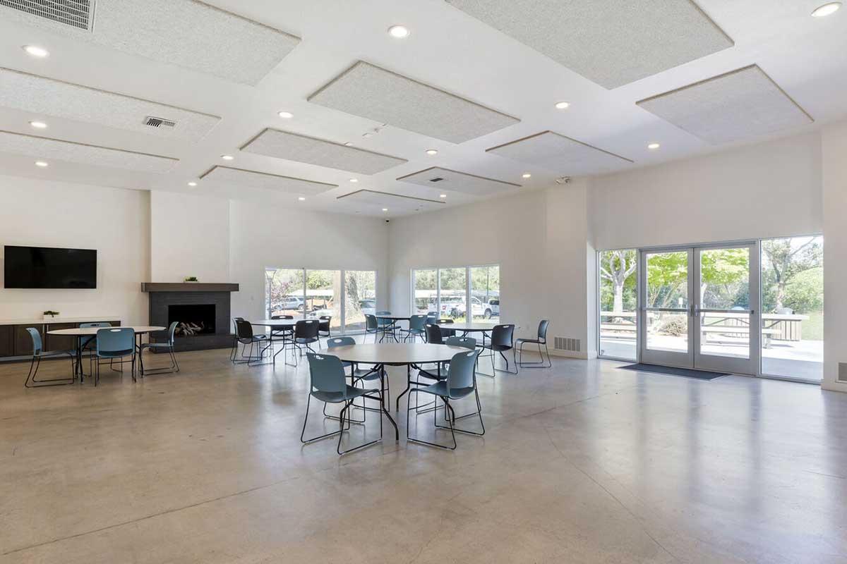 Spacious indoor gathering area featuring multiple round tables with chairs, large windows providing natural light, a fireplace, and a TV mounted on the wall.