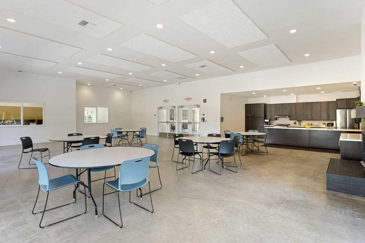 A spacious interior room with a modern design featuring several round tables, each surrounded by chairs. The flooring is concrete, and the walls are light-colored. One side of the room has a kitchen area with dark cabinets, a countertop, and appliances, while large windows allow natural light to enter.
