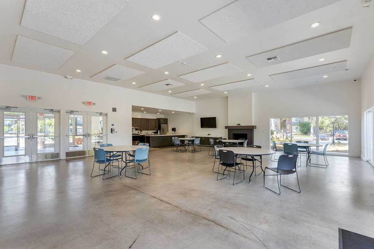 Interior of a spacious room with a modern design. Multiple round tables and chairs are arranged throughout the space. There is a kitchenette area on one side, a large TV mounted on the wall, and large windows letting in natural light. The flooring is concrete, and the ceiling features acoustic panels.