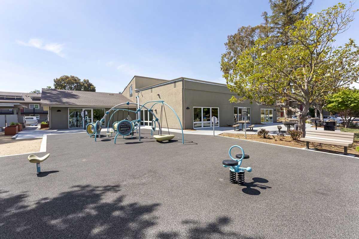 A playground area featuring swings, a spring rider, and a climbing structure, surrounded by a safety surface. In the background, a modern building with large windows and a landscaped area with trees.
