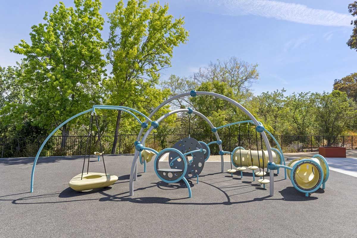 A modern playground structure featuring swings, a spinning disk, tunnels, and climbing elements, set against a backdrop of green trees and a clear blue sky. The playground equipment is designed for children to play and explore in a safe outdoor environment.