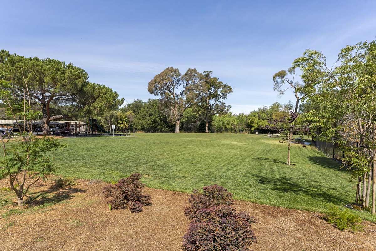 A spacious, green park area featuring a well-maintained grass lawn, surrounded by trees and shrubs. In the background, there are playground equipment and a fence, with a clear blue sky overhead. Ideal for outdoor activities and relaxation.