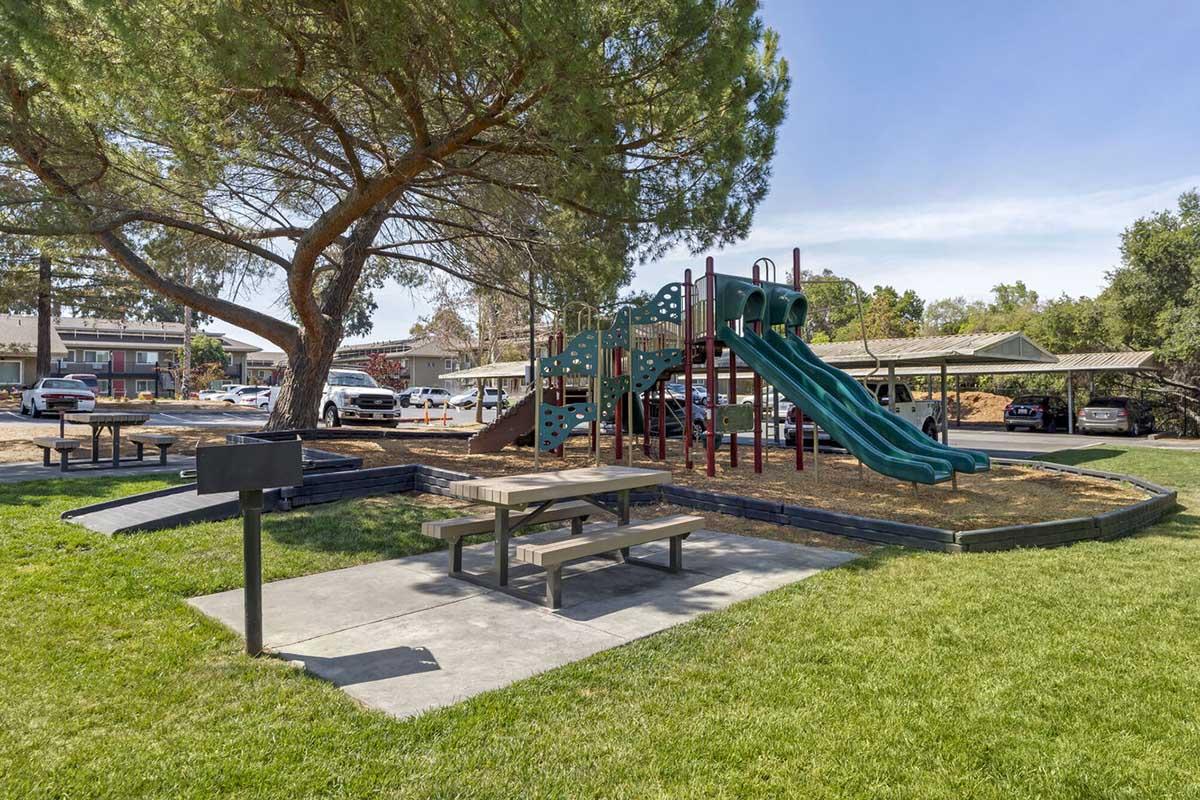 A playground featuring a colorful play structure with two green slides, surrounded by grass. Nearby, there are picnic tables under a large tree. In the background, parked cars are visible, along with a glimpse of nearby buildings. The scene is bright and inviting, perfect for outdoor play and relaxation.