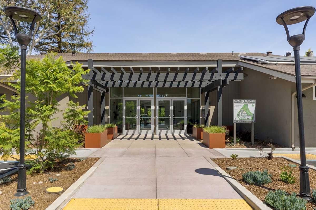 A modern building entrance with large glass doors, framed by a pergola. The pathway is lined with planters featuring green plants and shrubs.