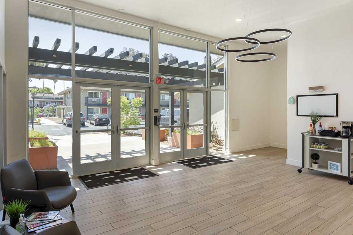 A spacious lobby area with large glass doors letting in natural light. It features modern lighting fixtures, a seating area with a black chair, and a small refreshments station with coffee. The view outside shows a landscaped patio and nearby buildings. The interior has neutral tones and wooden flooring.