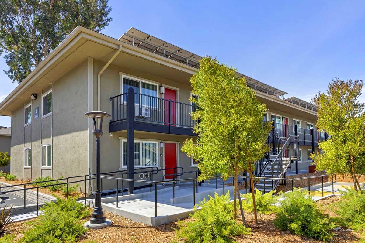 A modern two-story building with a gray exterior, featuring red doors and solar panels on the roof. The structure has a wheelchair-accessible ramp, and is surrounded by landscaped greenery and small trees. A pathway leads to the entrance, with a lamp post visible in the foreground.