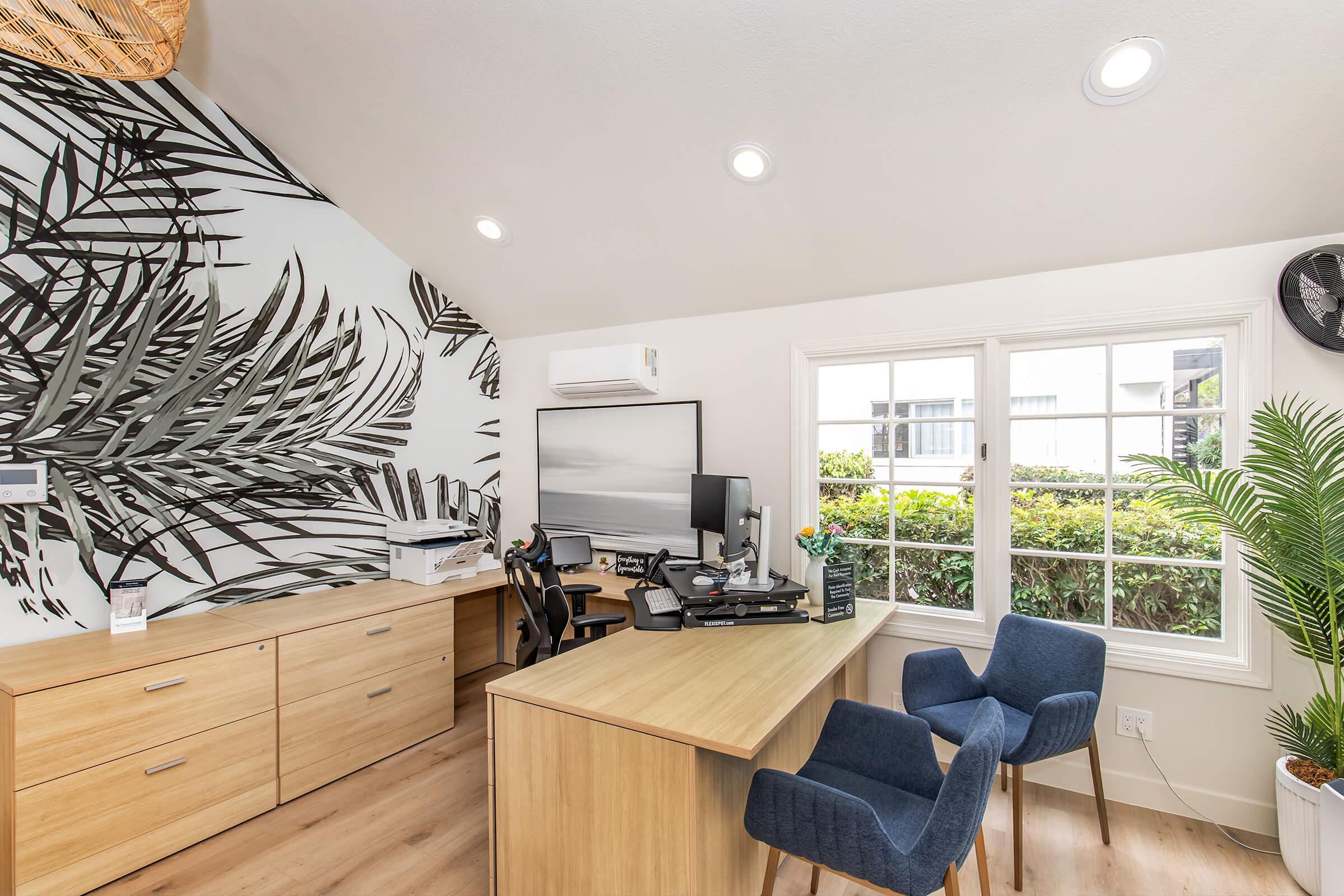 Modern home office featuring a wooden desk with a computer and office chair, surrounded by a tropical leaf mural on the wall. There is a printer on a cabinet, a potted plant near the window, and ample natural light illuminating the space. The overall design is bright and inviting.