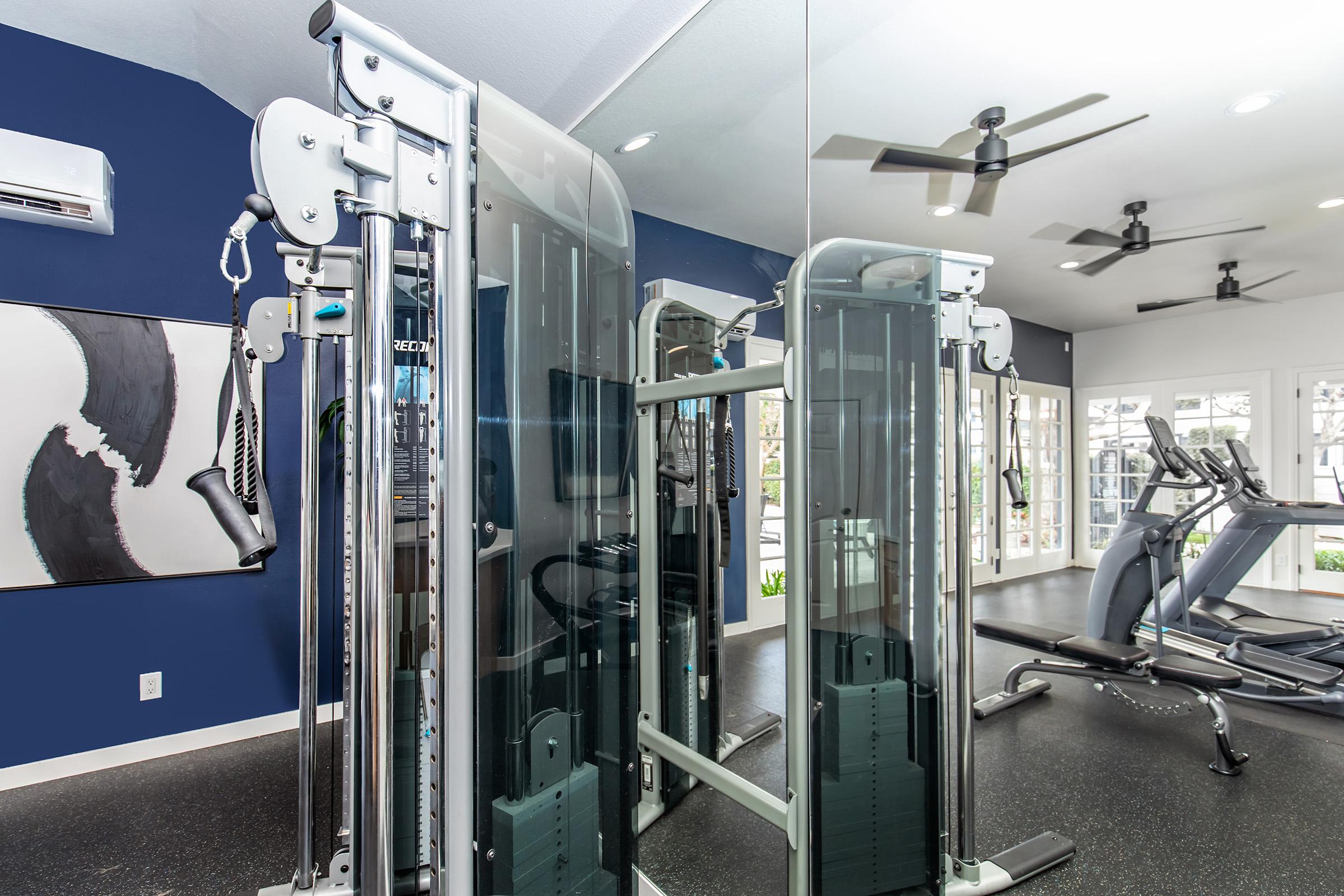 A modern gym with a cable machine in the foreground reflecting in a large mirror. The walls are painted dark blue, and there are exercise equipment and large windows allowing natural light. Ceiling fans are visible, enhancing the airy feel of the space.