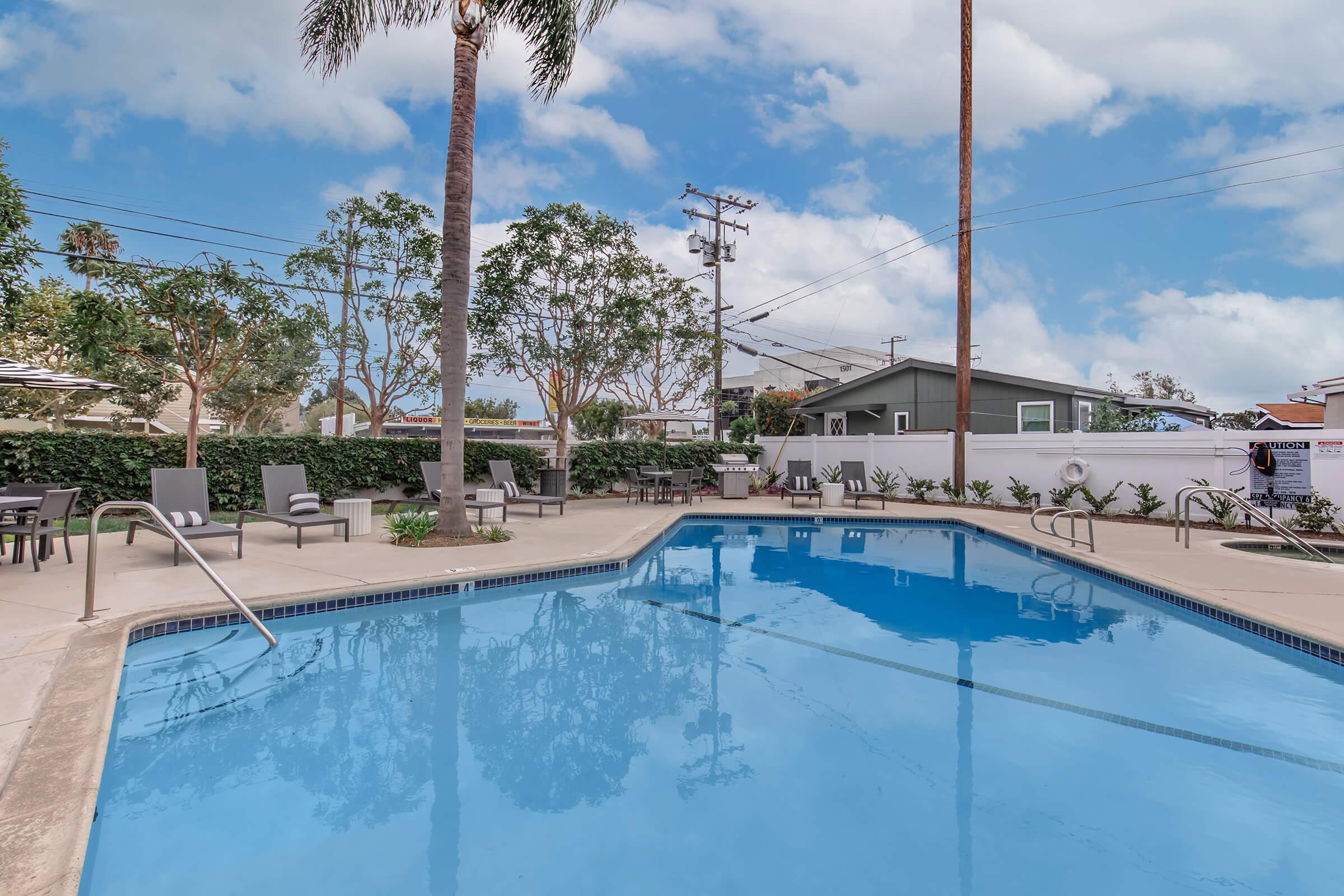 A clear blue swimming pool surrounded by lounge chairs and palm trees, with residential buildings and power lines in the background. The sky is partly cloudy, creating a bright and inviting atmosphere.