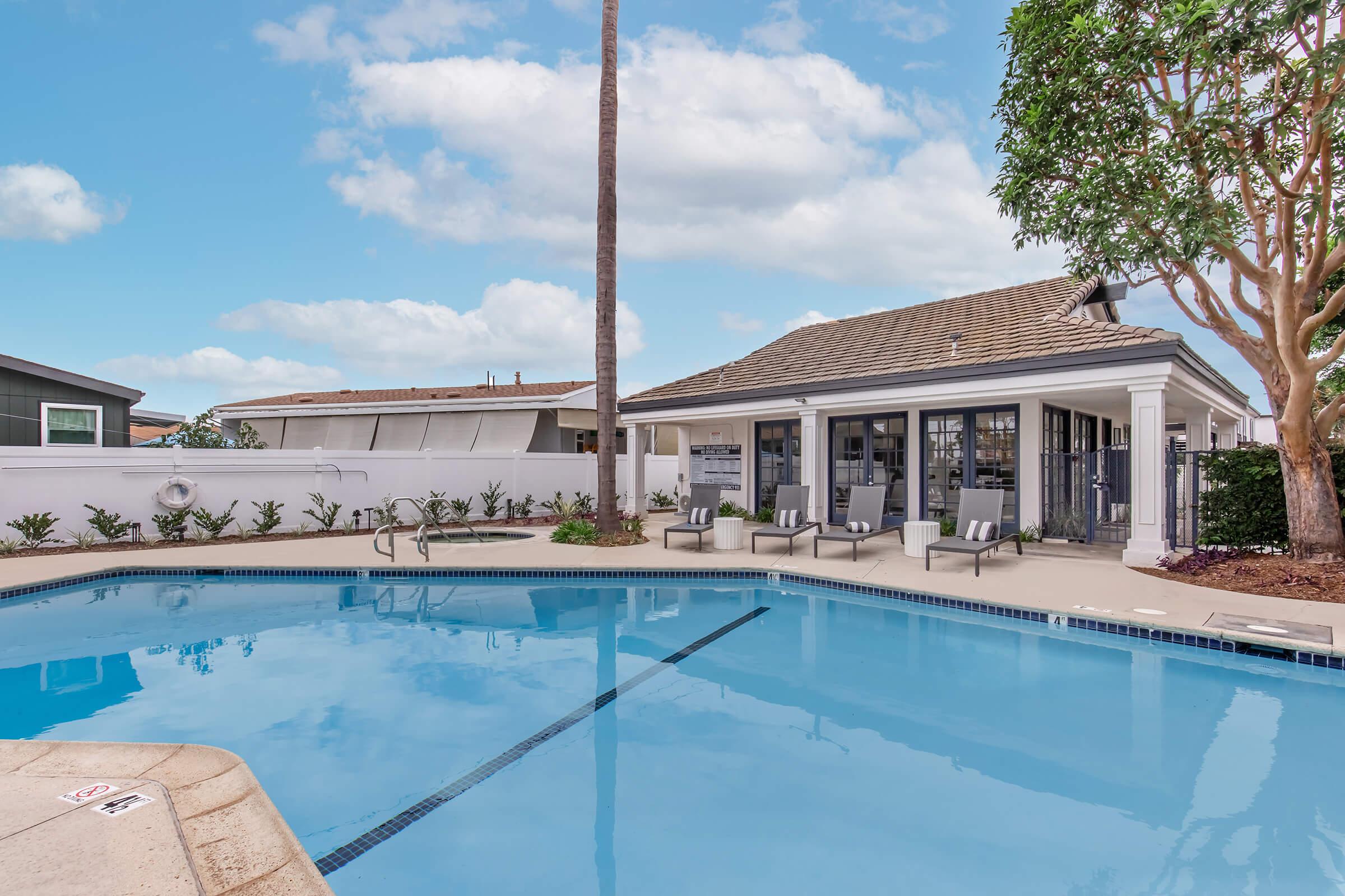 A clear blue swimming pool surrounded by lounge chairs and palm trees. In the background, there is a modern building with large windows and a patio area. The sky is partly cloudy, adding to a bright and inviting atmosphere.