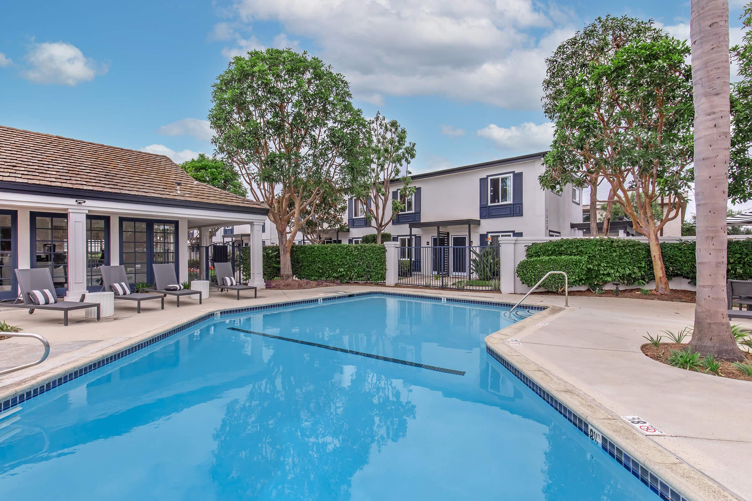 A serene swimming pool surrounded by lounge chairs and lush greenery. There are tall trees and a landscaped area with a fence in the background. The sky is clear with scattered clouds, creating a peaceful outdoor environment. Ideal for relaxation or leisure activities.