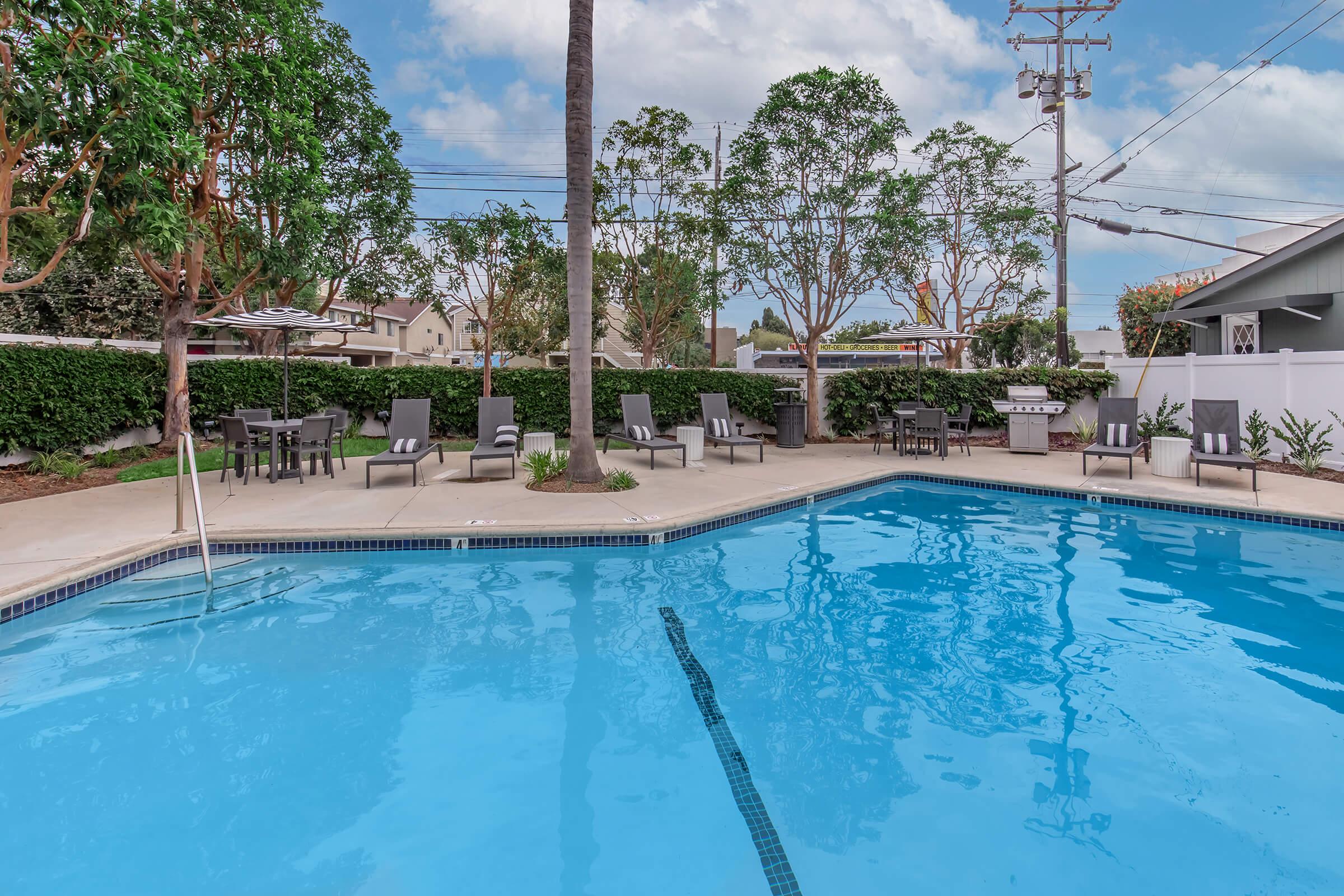 A clear blue swimming pool surrounded by lounge chairs and palm trees. The pool area features well-maintained landscaping with hedges and a grill, set against a backdrop of a sunny sky and residential buildings. Ideal for relaxation and outdoor gatherings.
