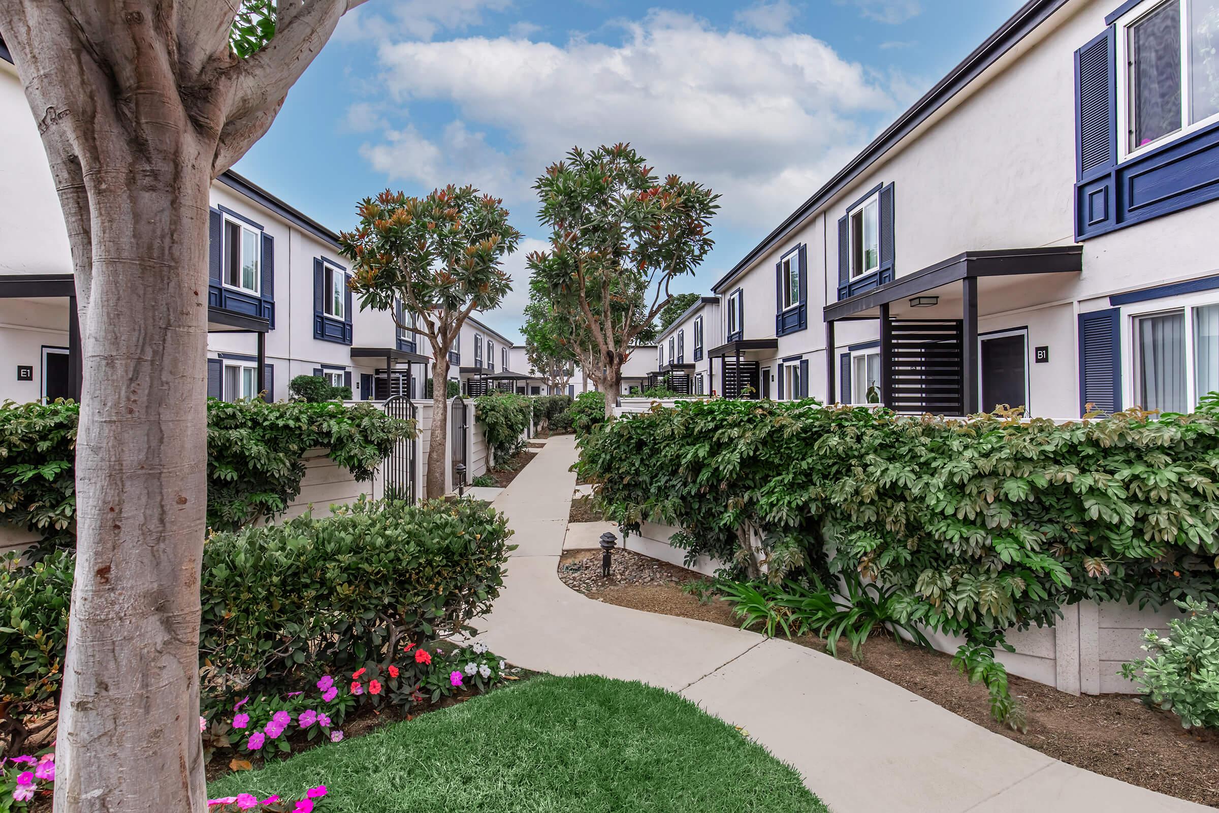 A well-maintained courtyard in a residential area, featuring a winding concrete pathway surrounded by lush greenery, flowering plants, and trees. Two-story buildings with blue accents line the pathway under a partly cloudy sky, creating a serene and inviting atmosphere.