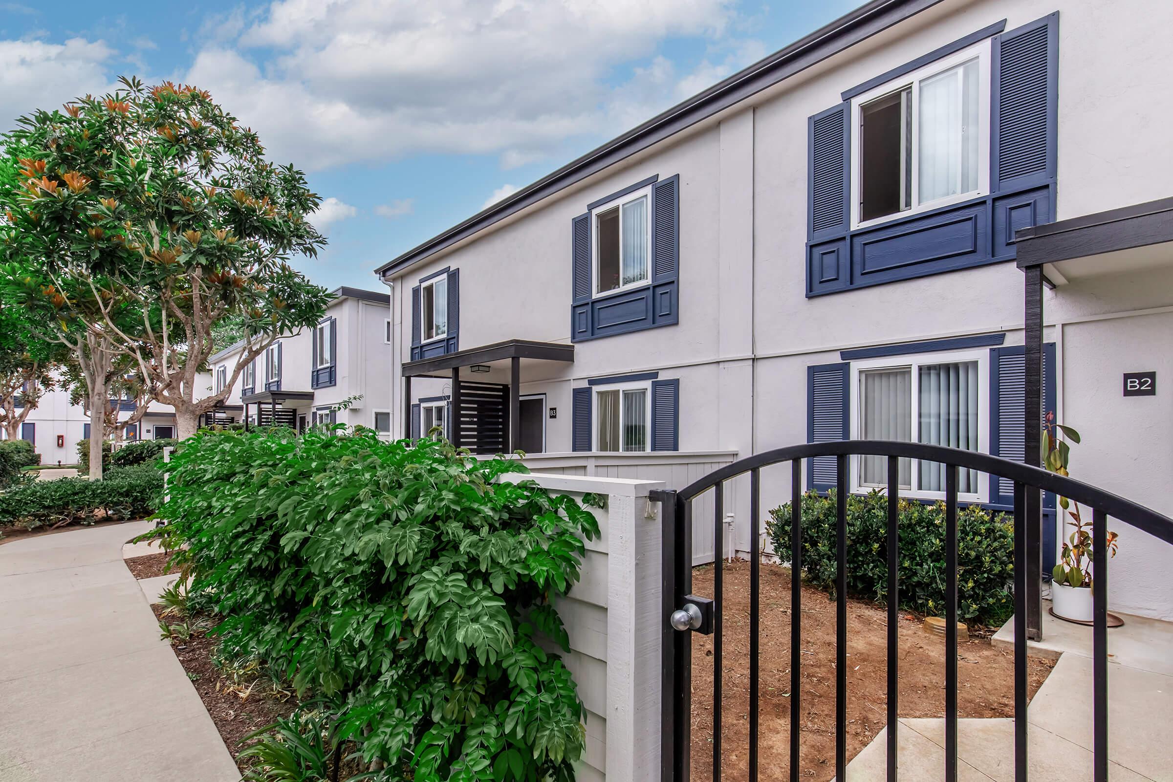 A view of a modern apartment complex featuring two-story buildings with blue shutters and white walls, surrounded by green landscaping. A gated entrance leads into a courtyard area with plants and pathways, under a partly cloudy sky.