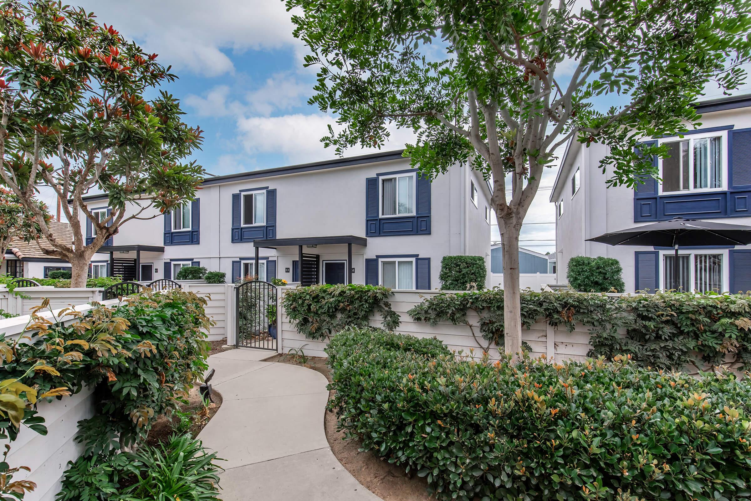 A landscaped pathway leads through lush greenery toward a modern building with blue accents and white walls. The scene features trees and shrubs, with a clear sky in the background, creating a welcoming and peaceful atmosphere.