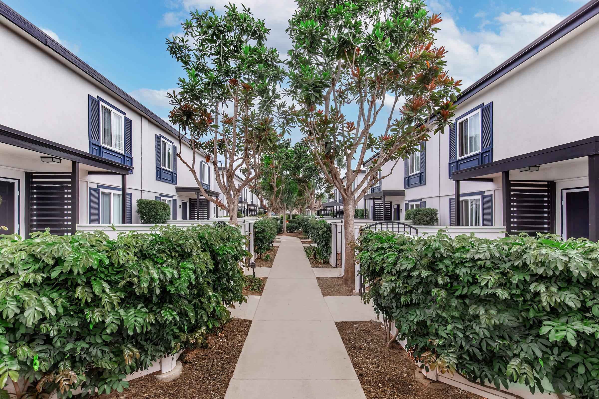 A landscaped pathway lined with shrubs leads through a residential area featuring two-story townhouse-like structures. The buildings have white exteriors and blue accents, with green trees providing shade. The sky is partly cloudy, creating a serene atmosphere.