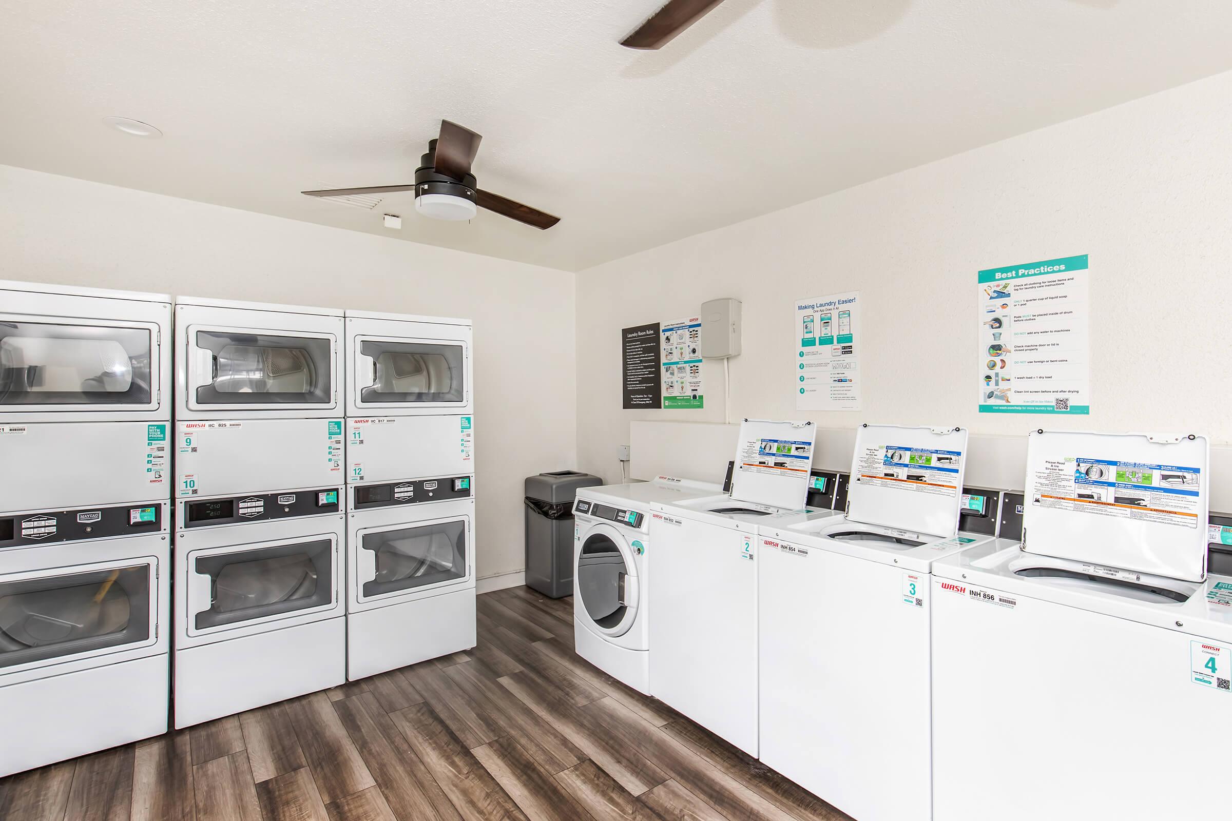 A clean laundry room featuring several white stacked washers and dryers. The room has a ceiling fan, and the walls are adorned with instructional signs and posters about laundry best practices. The flooring is made of dark wood planks, enhancing the modern and tidy atmosphere.