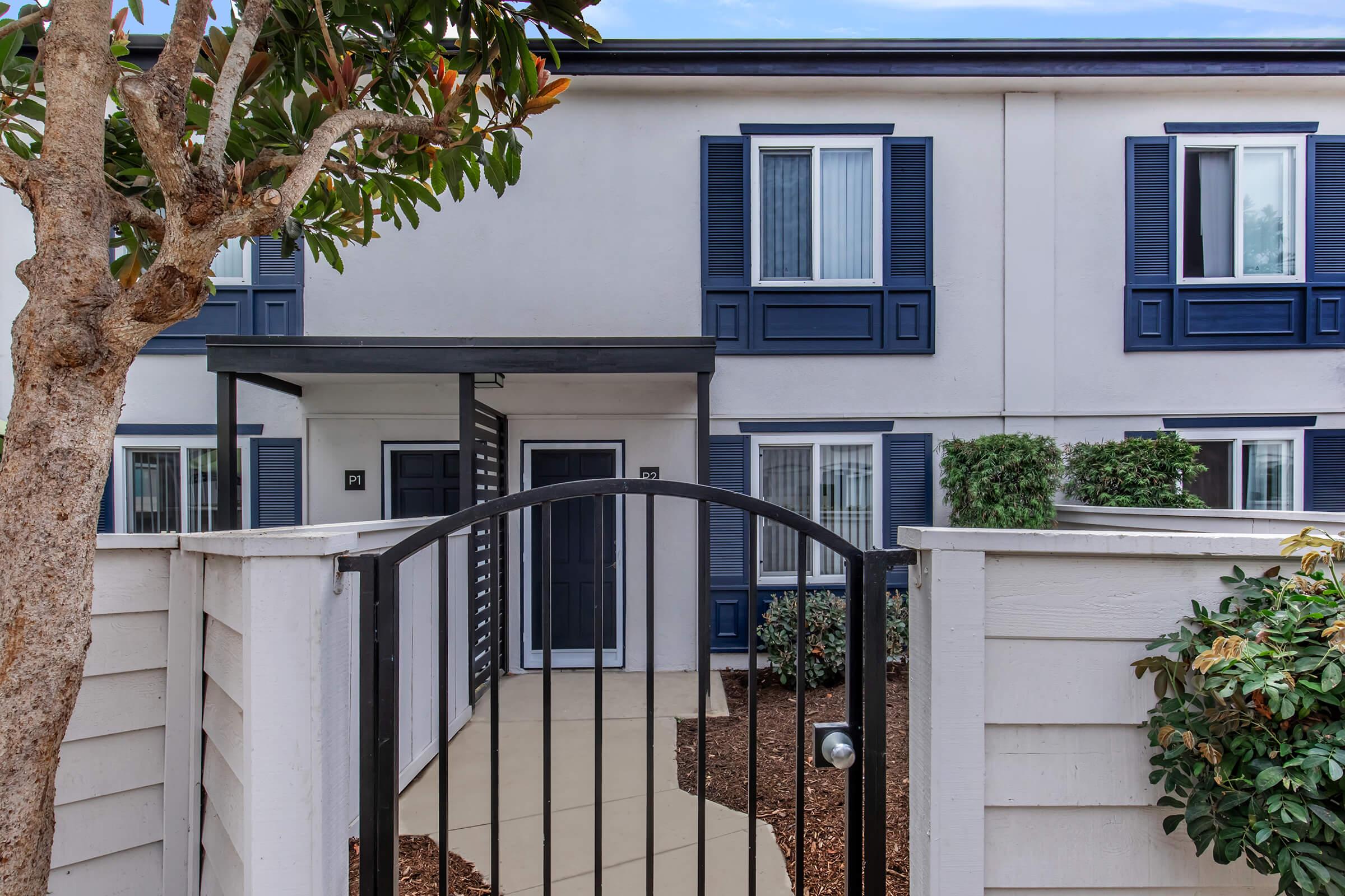 A modern building entrance featuring a gated pathway. The facade has white walls with blue shutters on the windows, a covered porch area, and well-maintained landscaping with shrubs and small trees flanking the entrance. The door is framed by a simple, elegant design, inviting visitors in.