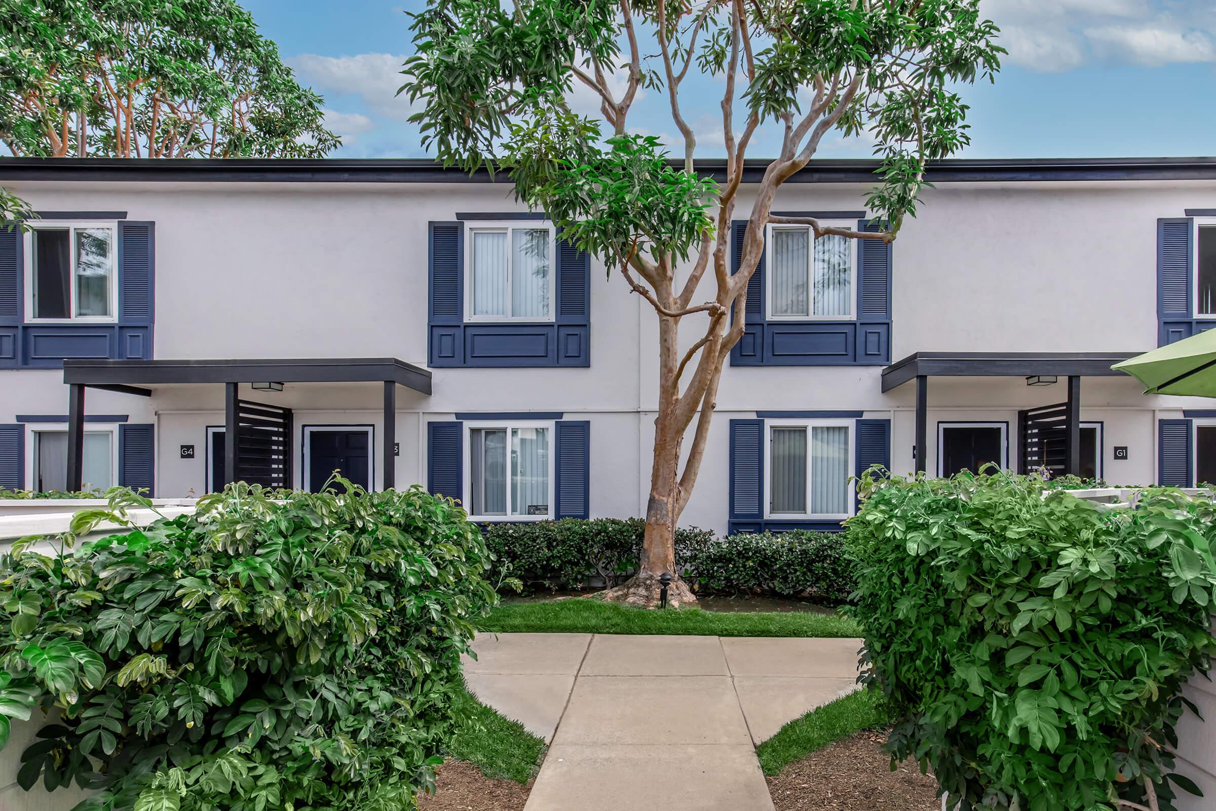 A landscaped pathway leads to a two-story building with multiple windowed units, surrounded by green shrubbery and trees. The facade features blue shutters and a modern design under a bright sky.