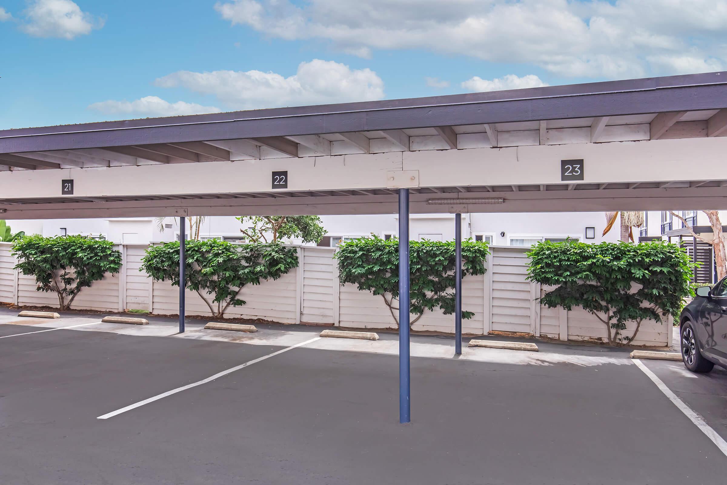 A parking lot with two covered spaces numbered 22 and 23. The area has a neatly maintained light-colored fence with green shrubs on either side. The sky is clear with scattered clouds, creating a bright and open atmosphere.