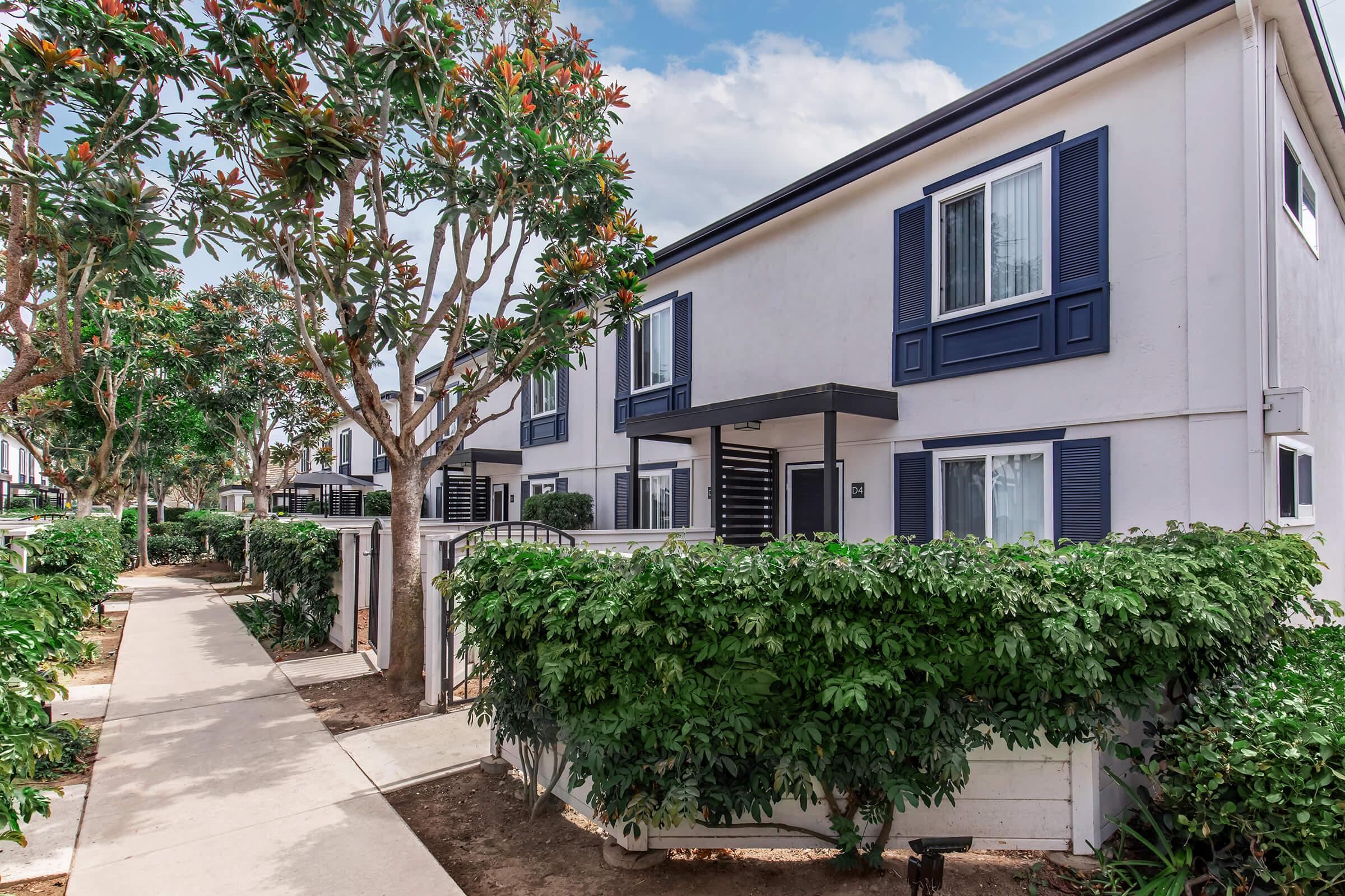 A pathway lined with lush greenery leads to a row of modern two-story residential buildings. Each unit features blue shutters and large windows, surrounded by well-maintained landscaping, creating a welcoming atmosphere in a tranquil neighborhood.