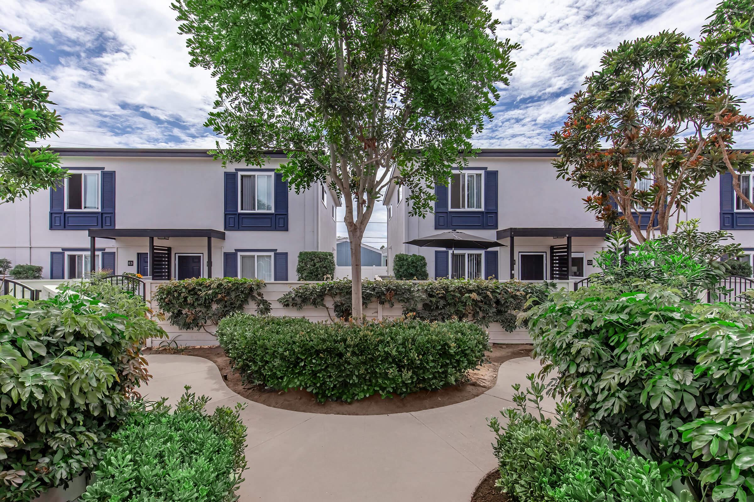A landscaped courtyard with lush greenery and a circular pathway leads to two modern buildings featuring blue accents. The sky is partly cloudy, creating a bright atmosphere, while trees provide shade and ambiance to the area.