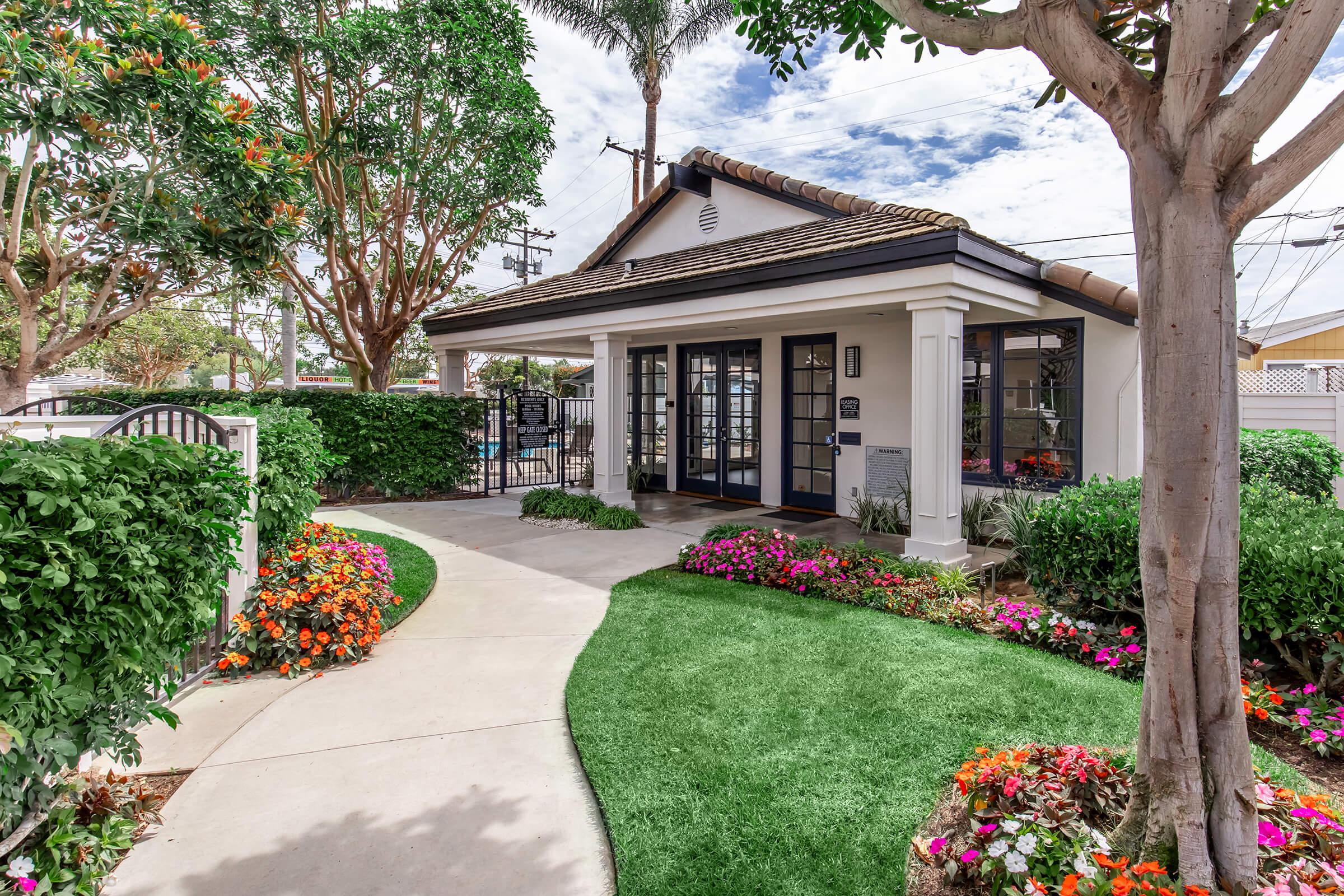 A charming residential entrance with a landscaped walkway leading to a house. Lush green lawn and vibrant flower beds frame the path. The house features a light-colored exterior with dark window frames and a tiled roof, surrounded by trees and shrubs under a partly cloudy sky.