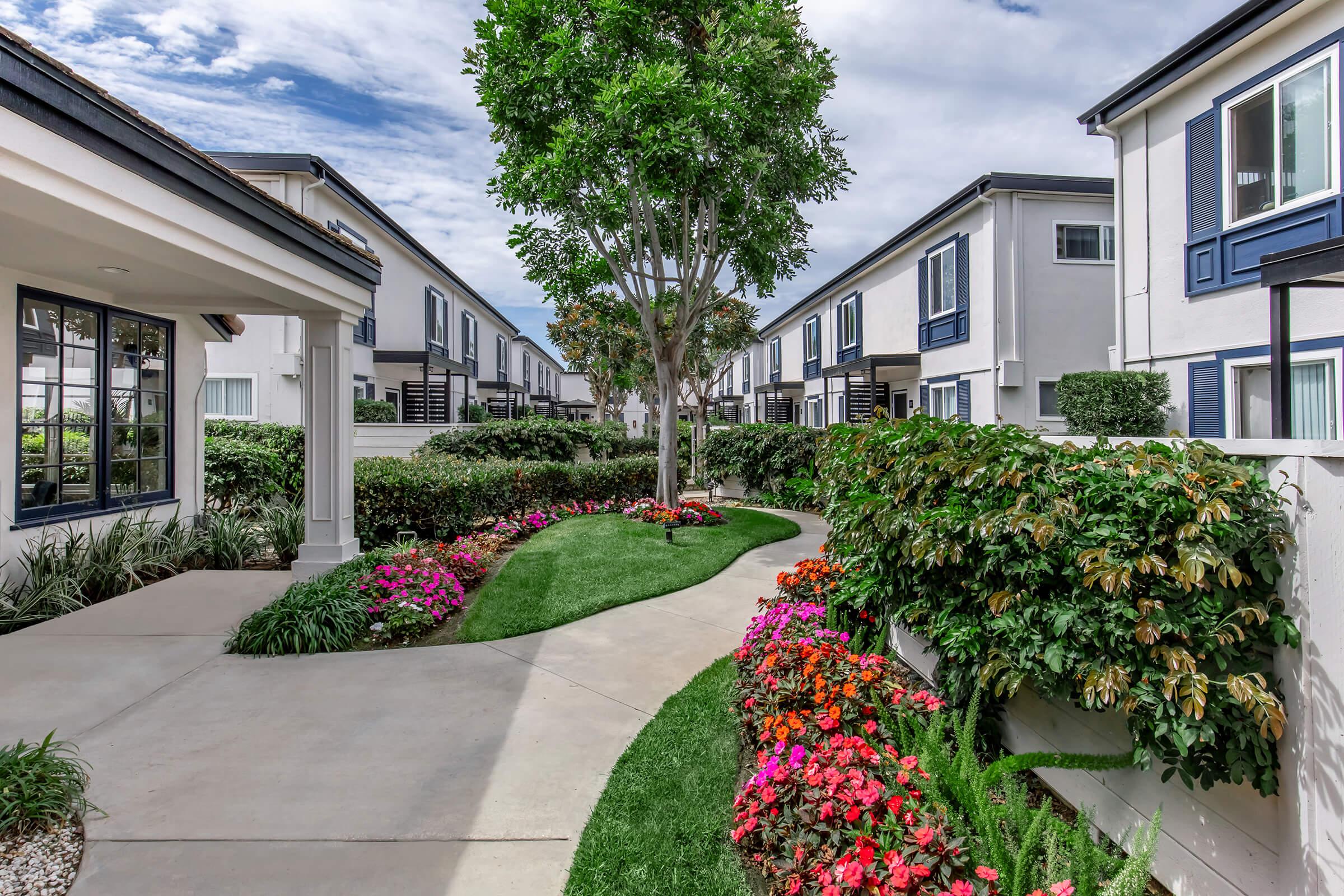 A landscaped pathway lined with colorful flower beds leads through a modern residential area. Lush green grass and trees enhance the aesthetic, while contemporary buildings with blue accents form the backdrop under a partly cloudy sky.