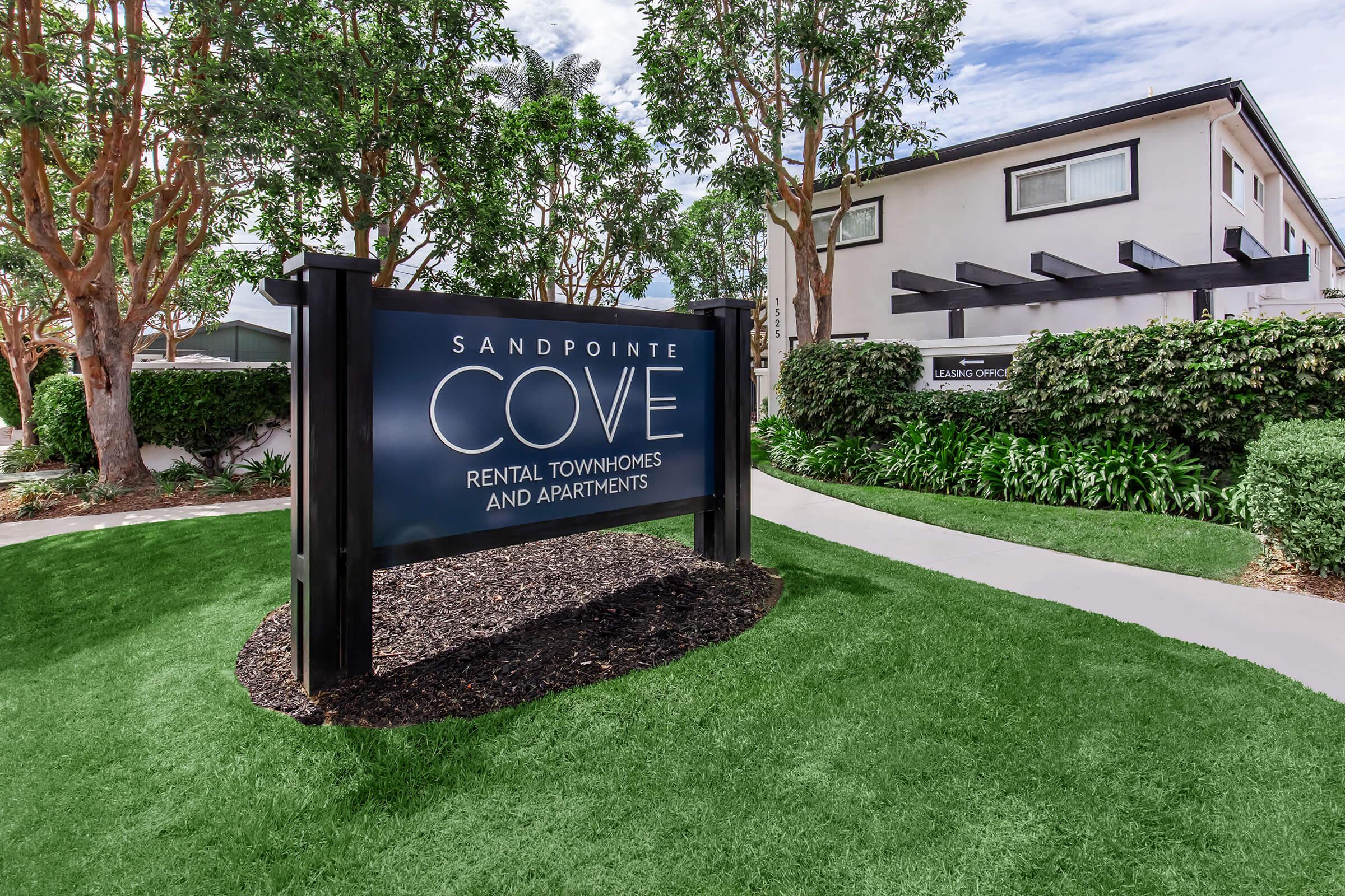 Sign for Sandpointe Cove featuring the text "Sandpointe Cove" and "Rental Townhomes and Apartments." The sign is surrounded by lush greenery and well-maintained landscaping, with a pathway leading towards a modern building in the background. The sky is partly cloudy, enhancing the inviting atmosphere of the location.