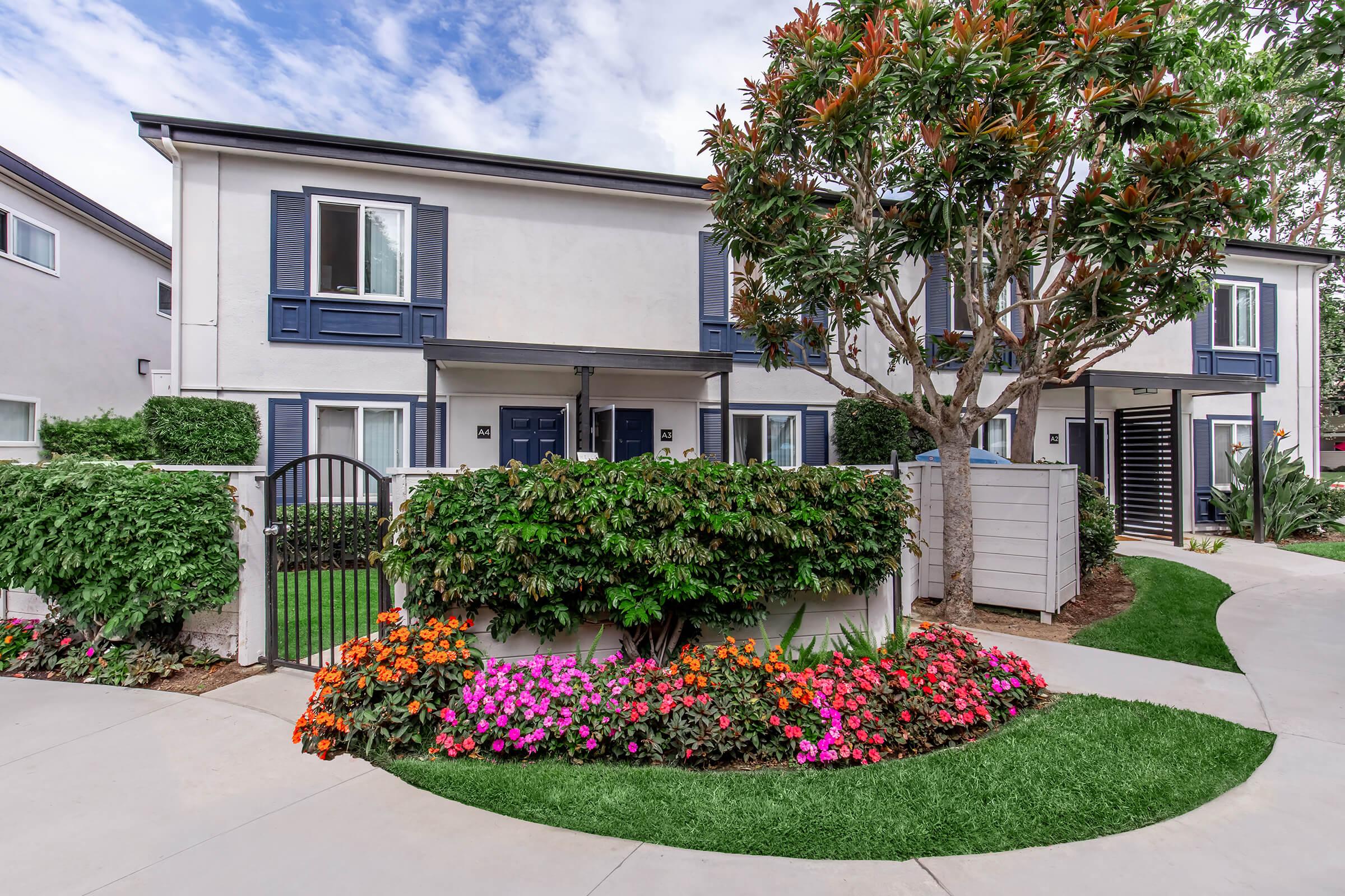 A well-maintained residential building with blue shutters and a landscaped garden featuring colorful flowers. A curved walkway leads to the entrance, bordered by lush greenery. A tree stands prominently in the garden, enhancing the inviting atmosphere of the property.
