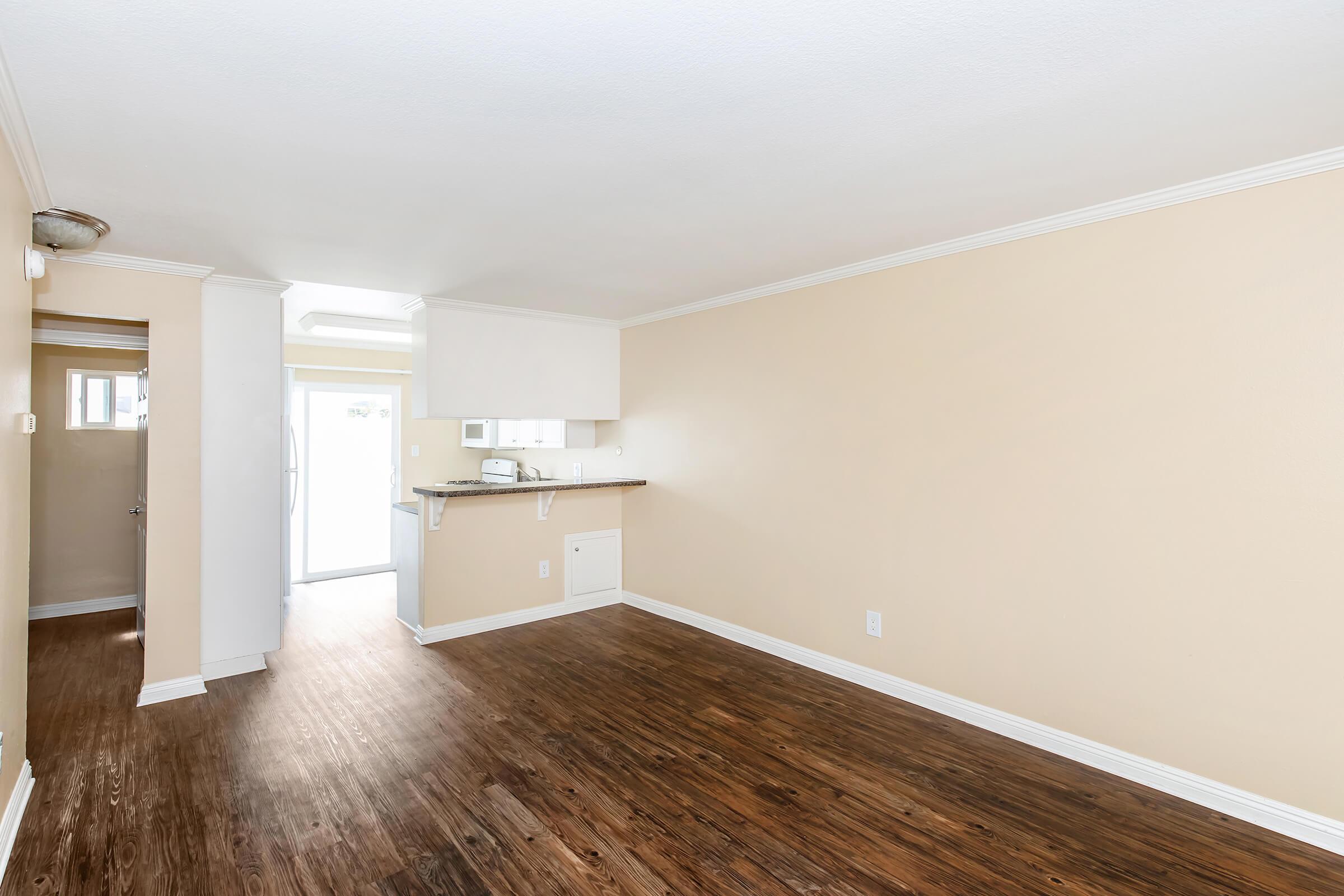 A spacious, unfurnished room with light beige walls and a wood floor. There is a simple kitchen area on the right with a countertop, and a door leading outside. Natural light enters from the door, giving the room a bright and airy feel. The room is ready for personal decoration and furniture.