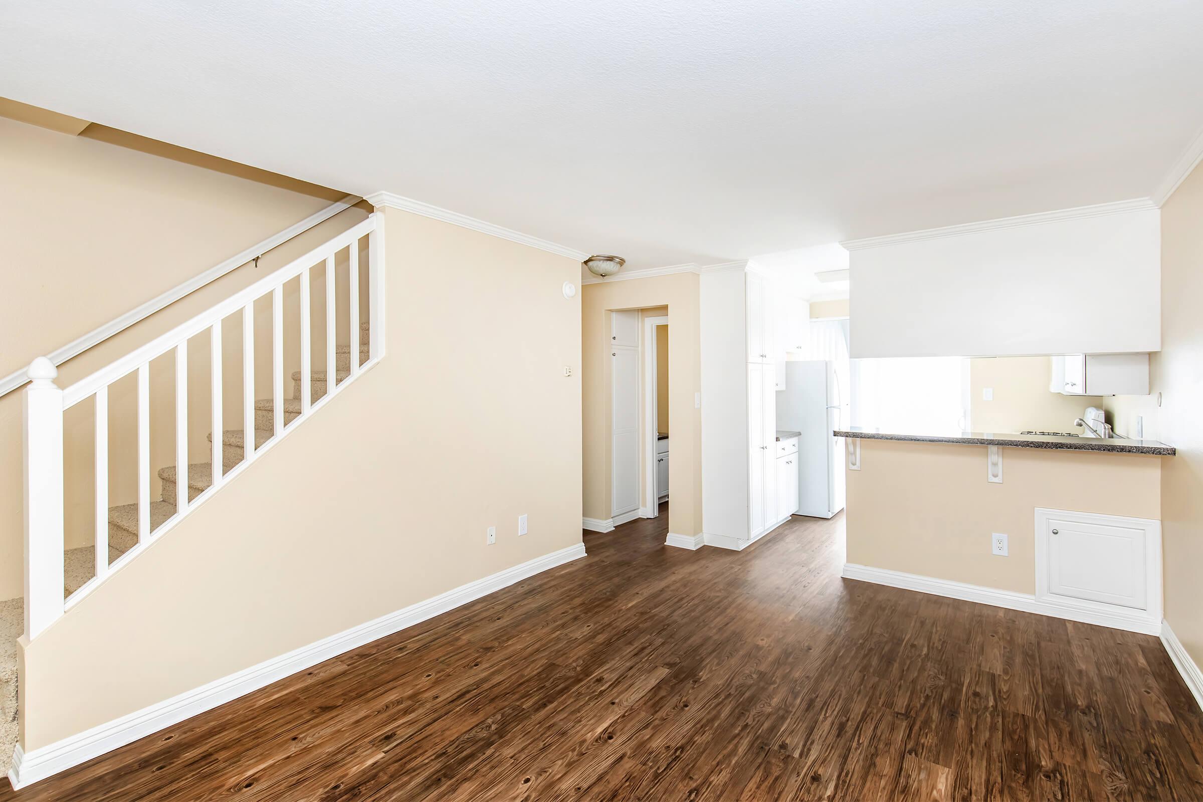 Bright, open-concept living space featuring a wooden floor, light-colored walls, and a staircase in the background. The kitchen is visible with a white refrigerator and countertop. Natural light illuminates the area, creating a warm and inviting atmosphere. Spacious and ideal for various furnishings.