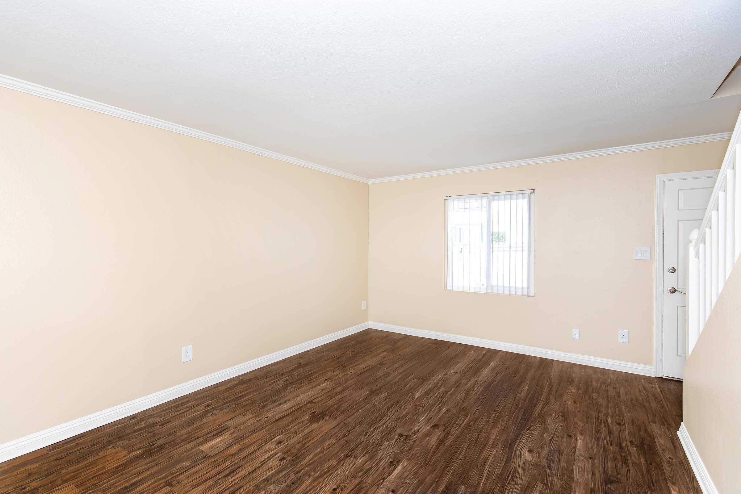 A spacious and empty room with light beige walls and wooden laminate flooring. A window with vertical blinds allows natural light to enter, and there's a door leading outside. The ceiling has white molding, enhancing the room's clean and airy appearance.