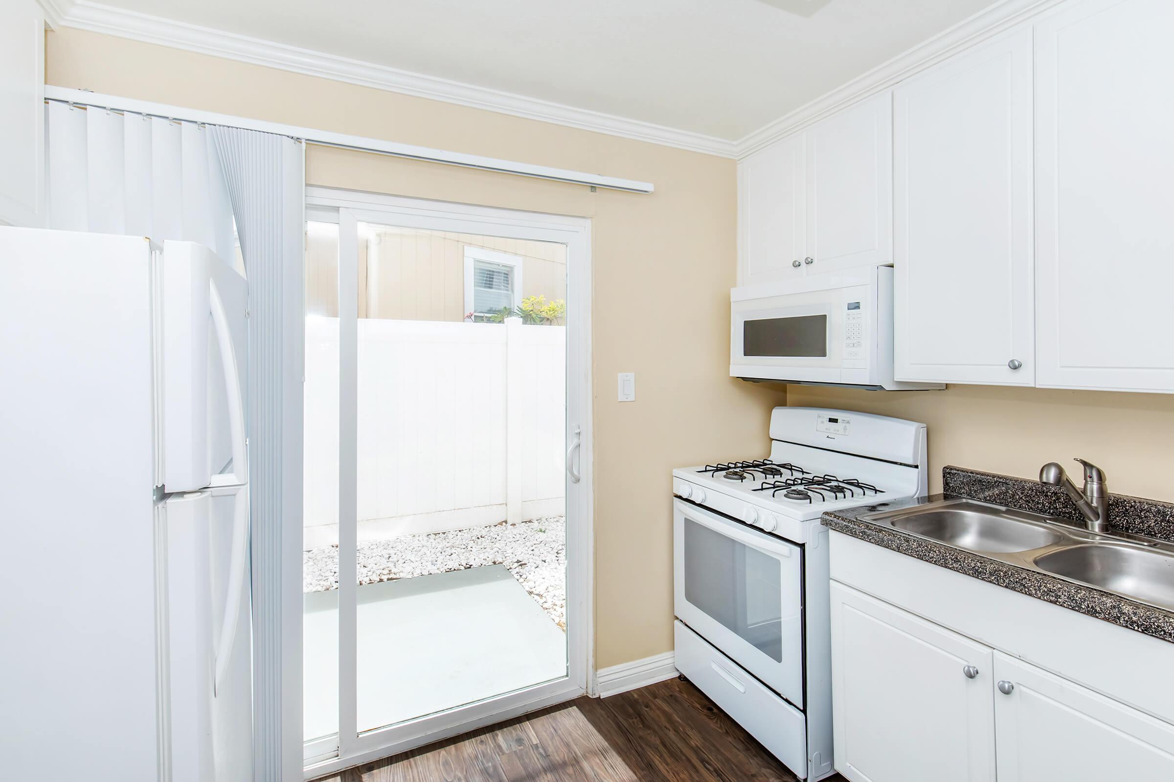 A bright kitchen with white cabinetry and appliances, featuring a stove, microwave, and sink. A window above the sink overlooks a small outdoor area. The floor has a wood-like finish, and there's a sliding glass door leading to the outside.