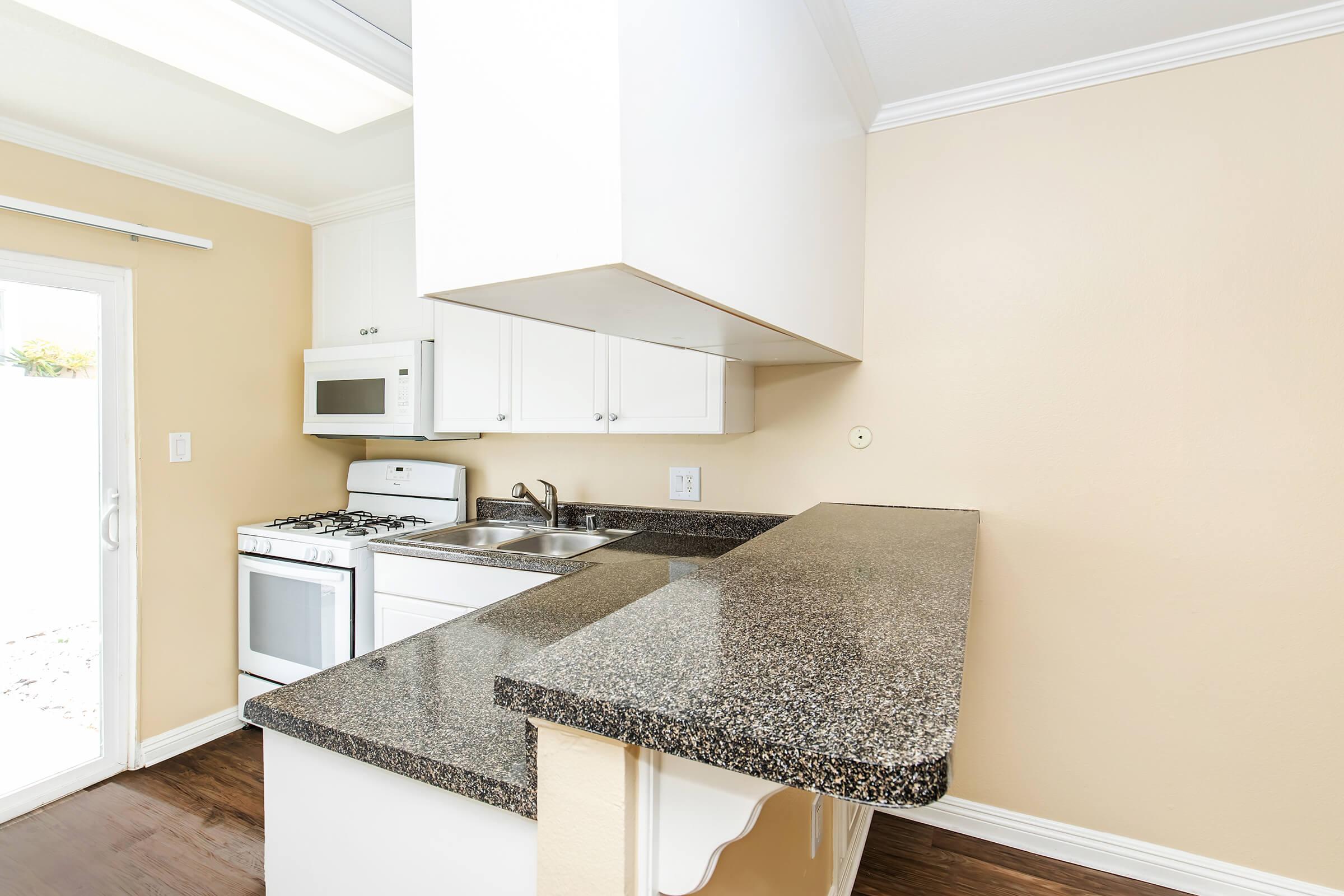 A modern kitchen featuring white cabinets, a granite countertop, an oven, a microwave, and a sink. Natural light enters through a nearby window, highlighting the warm beige walls and dark wood flooring. The design is minimalist and clean, suitable for a small space.