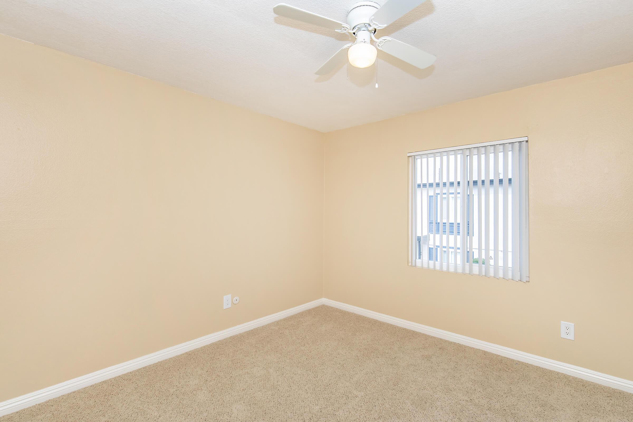 Empty room with light beige walls and a ceiling fan. A window with white blinds lets in natural light. The floor is covered with light brown carpet, creating a simple and clean atmosphere.