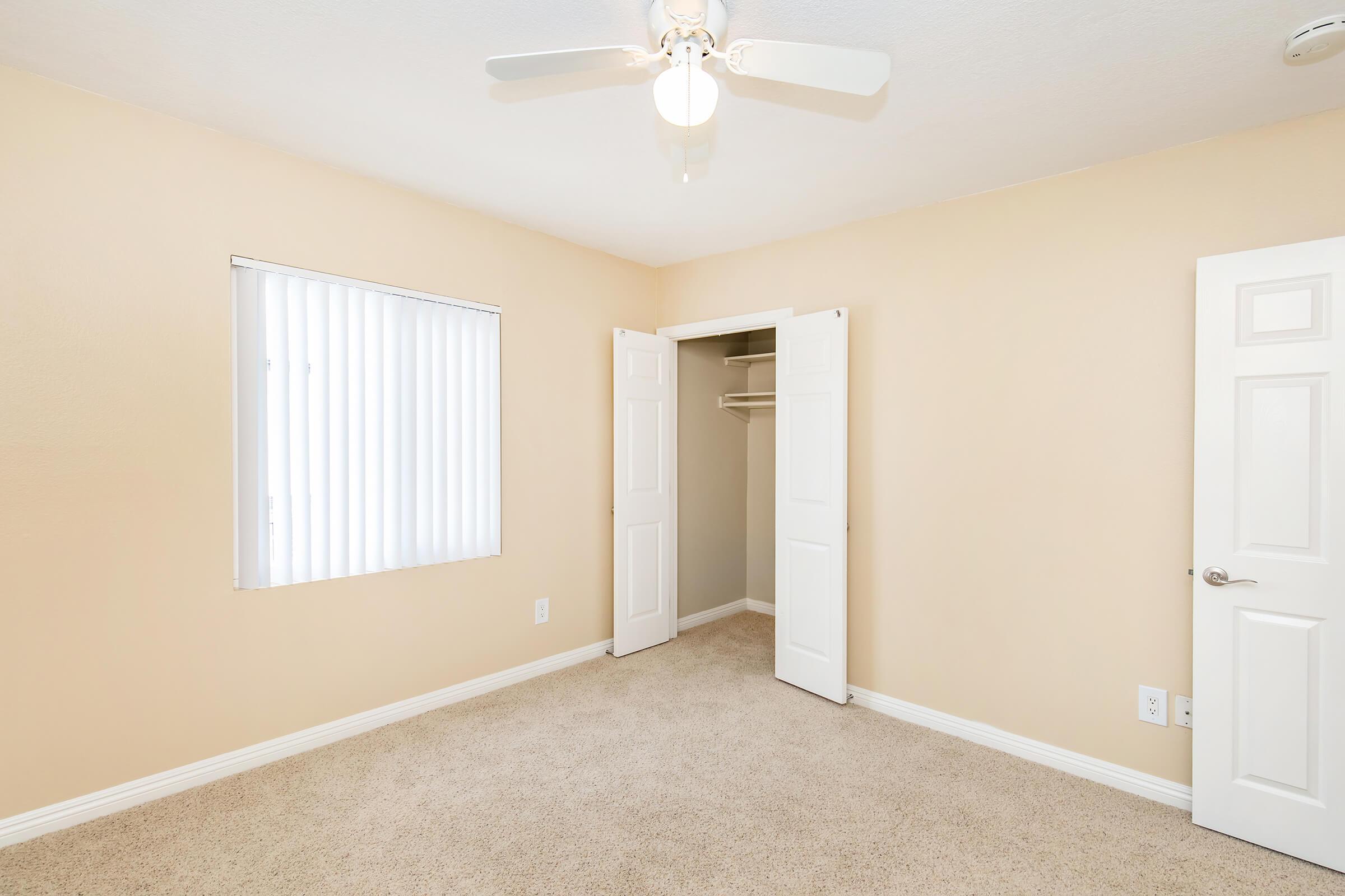 Empty room with beige walls and carpet flooring. A ceiling fan is installed on the ceiling, and a window with vertical blinds allows natural light. A closet with double doors is open, revealing a simple, uncluttered interior. The overall ambiance is clean and bright, ideal for personal decoration.